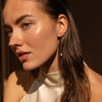 A woman with dewy skin and wet hair glows in soft sunlight, wearing minimalistic Pearl Threader Earrings and a cream halter top.