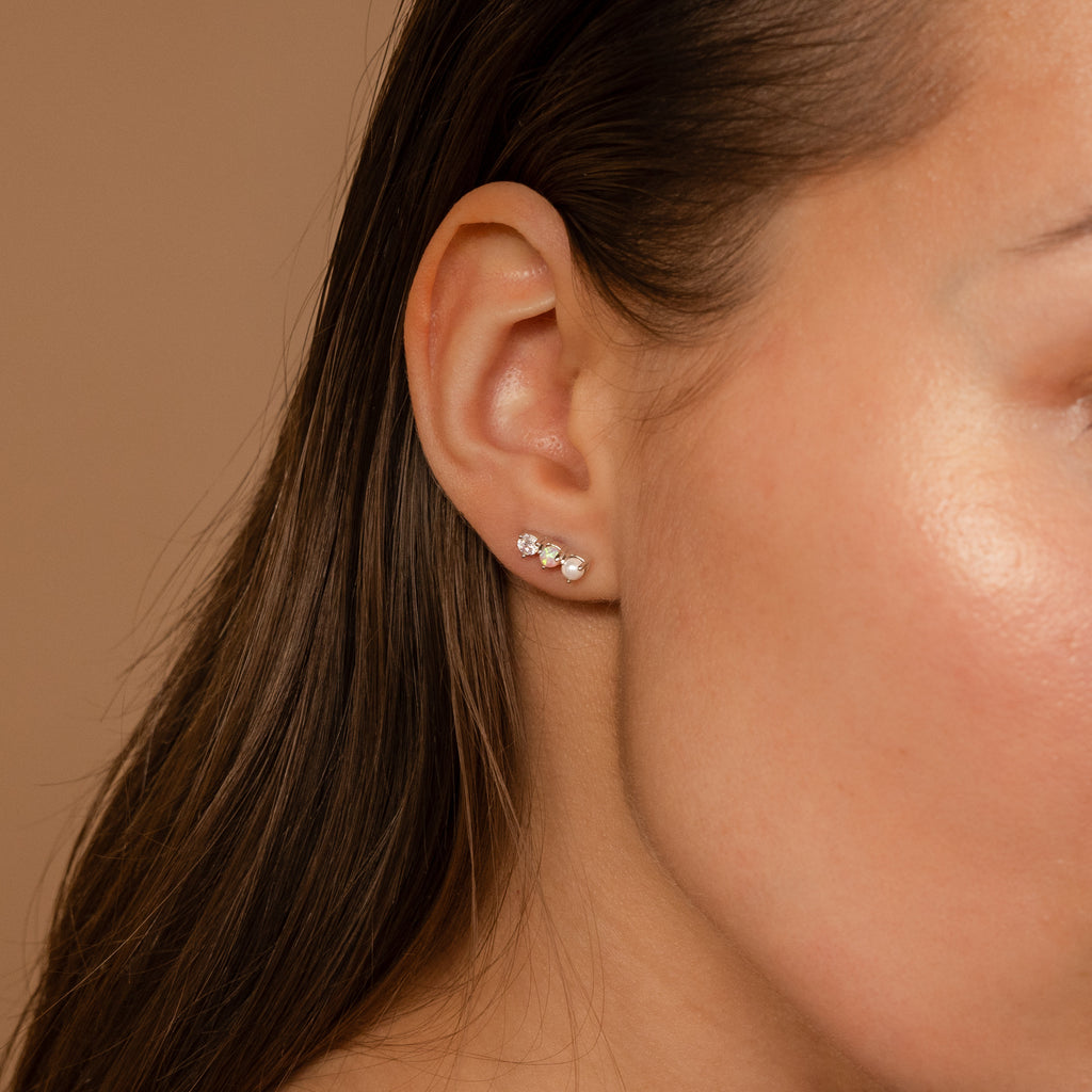 A close-up of a woman's ear wearing the Pearl, Opal & Diamond Studs set—three small, elegant earrings—against a neutral background.
