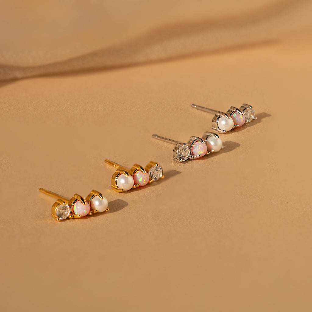 Three pairs of Pearl, Opal & Diamond Studs in gold, silver, and rose gold are displayed on a beige surface.