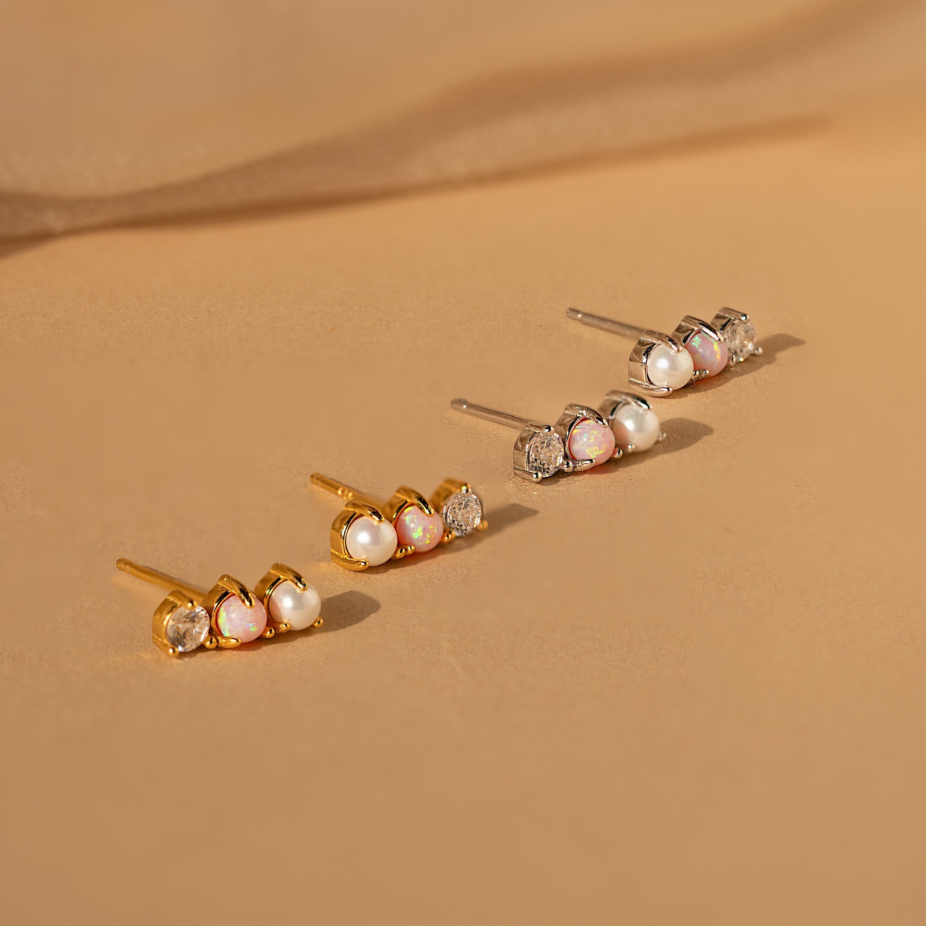 Three pairs of Pearl, Opal & Diamond Studs in gold, silver, and rose gold are displayed on a beige surface.