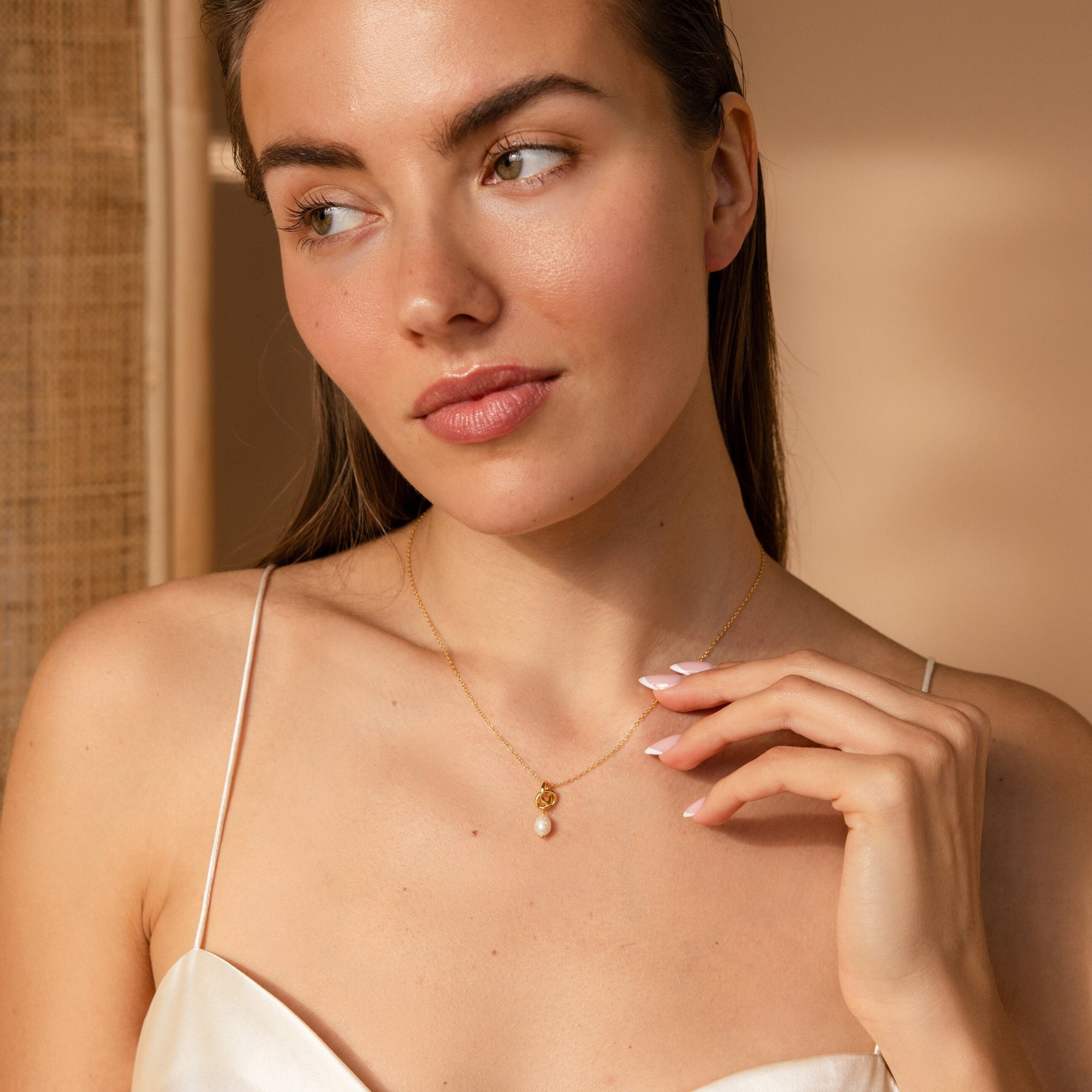 A woman in a white top gently touches the pearl charm of the Dainty Knot Pearl Necklace and gazes to the side.