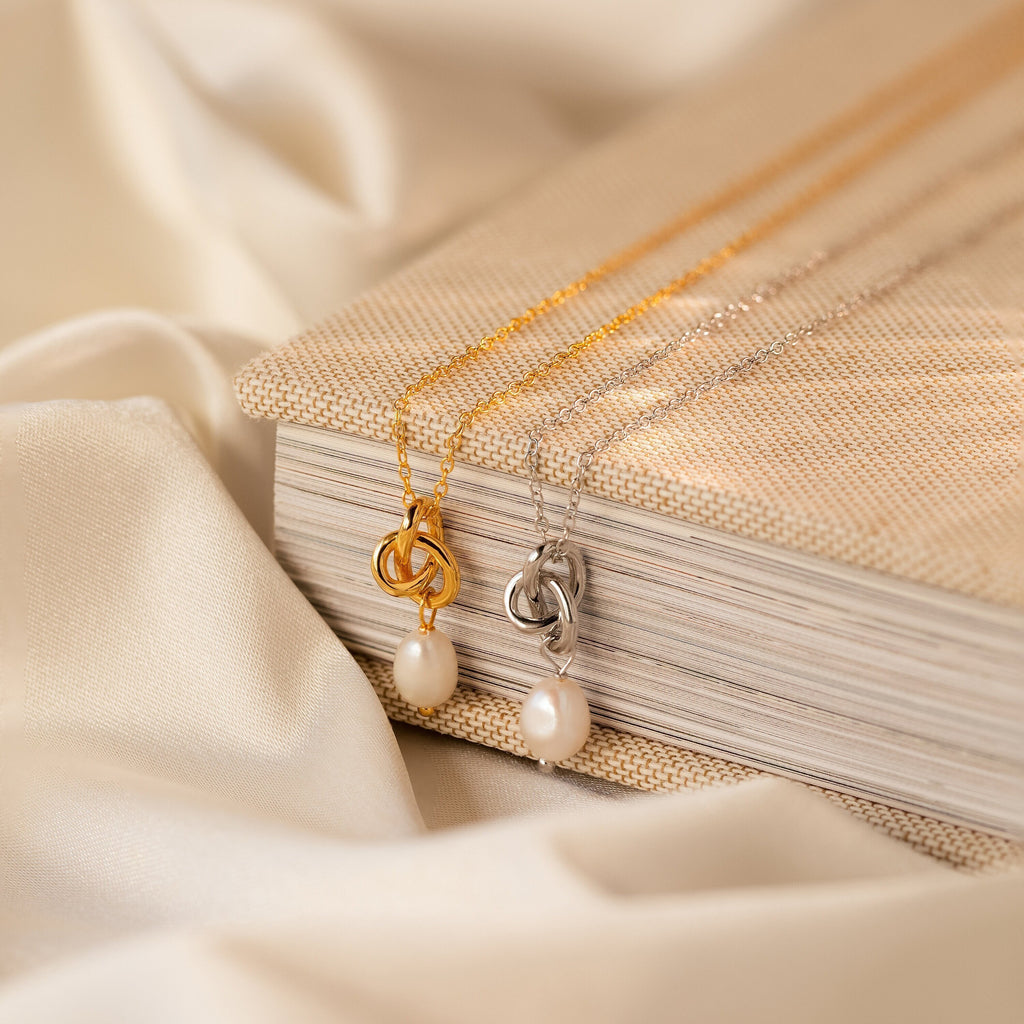 The Dainty Knot Pearl Necklace is displayed on a textured beige book, surrounded by soft, cream-colored fabric.