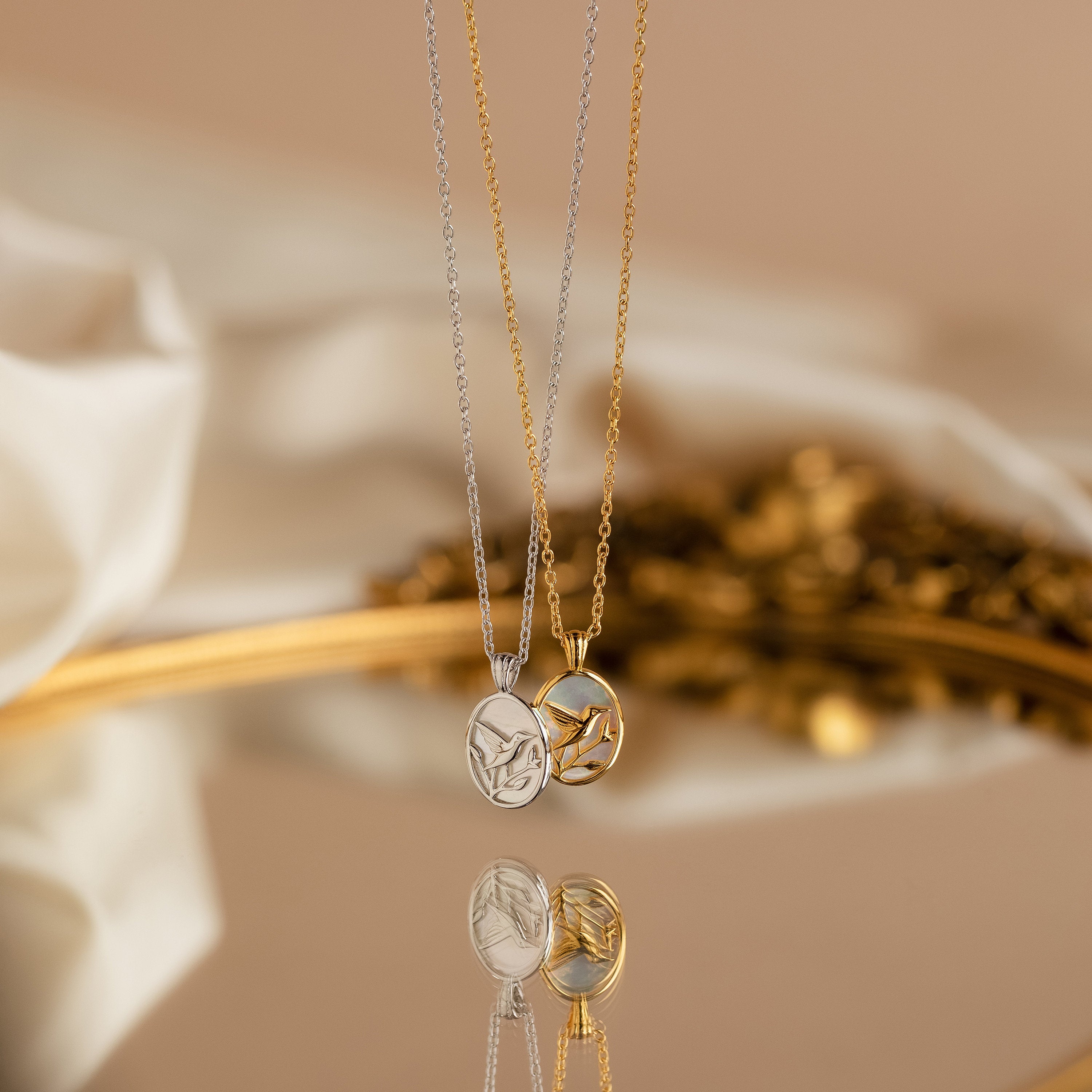 Two Pearl Hummingbird Necklaces, one silver and one gold with round pendants, are displayed on a reflective surface—ideal for those seeking a minimalist necklace with vintage-inspired charm.