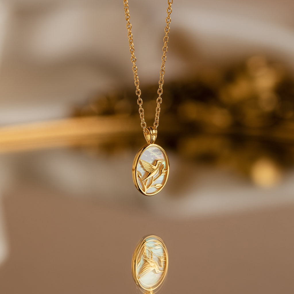 The Pearl Hummingbird Necklace in 18K Gold features an elegant bird design on an oval charm, beautifully reflected on a shiny surface. This vintage-inspired piece adds timeless sophistication to any look.