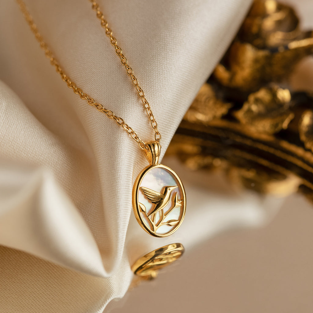 The Pearl Hummingbird Necklace features an oval pendant with a hummingbird design, displayed on soft cream fabric—a vintage-inspired, minimalist gold necklace perfect for subtle elegance.