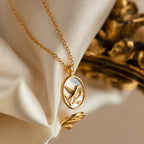 The Pearl Hummingbird Necklace features an oval pendant with a hummingbird design, displayed on soft cream fabric—a vintage-inspired, minimalist gold necklace perfect for subtle elegance.