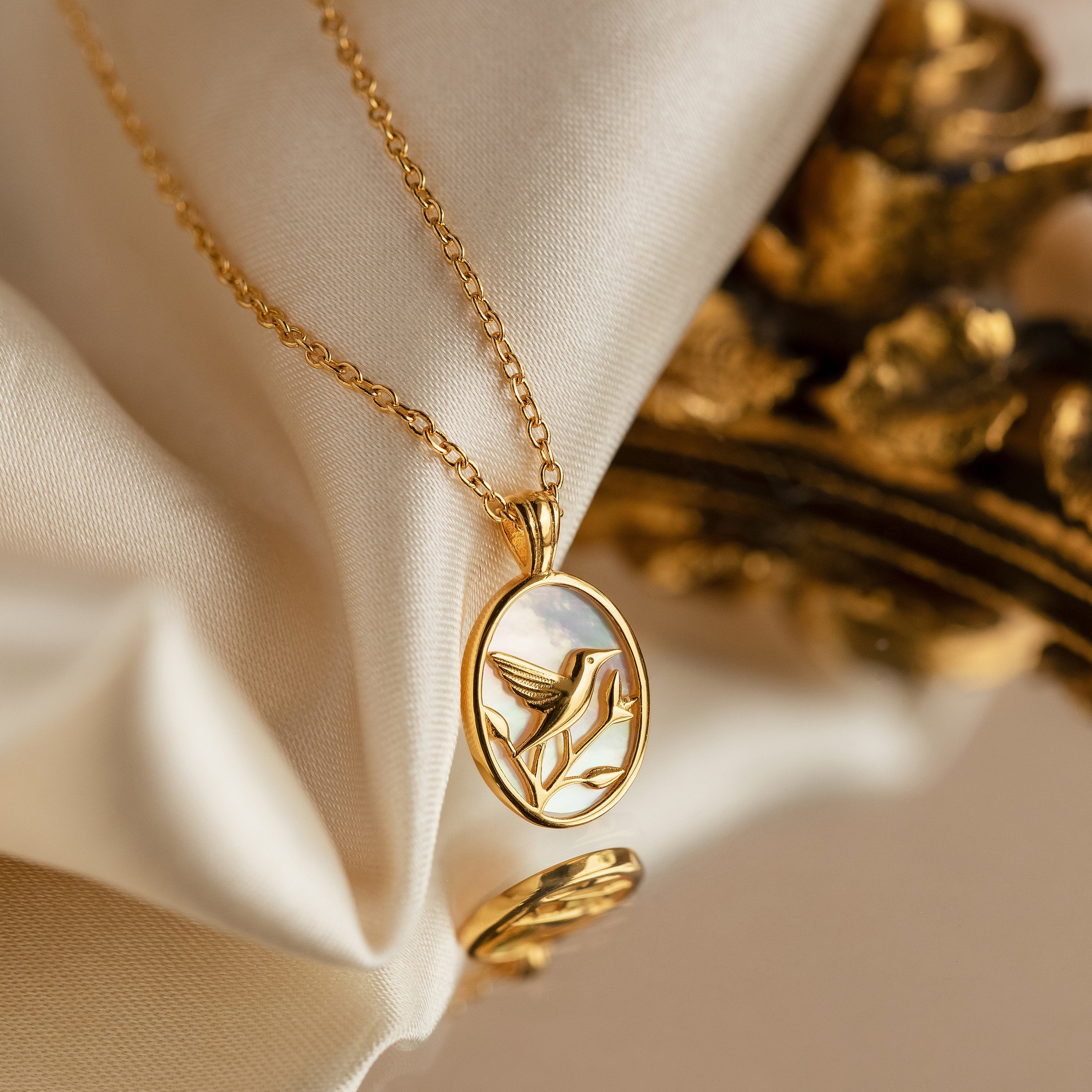 The Pearl Hummingbird Necklace features an oval pendant with a hummingbird design, displayed on soft cream fabric—a vintage-inspired, minimalist gold necklace perfect for subtle elegance.