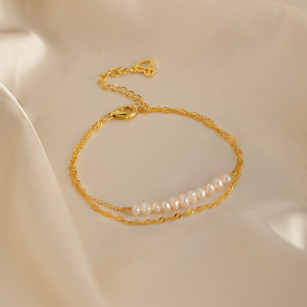 The Duo Pearl Twist Bracelet, featuring gold accents and white pearls, is elegantly displayed on soft beige satin fabric.