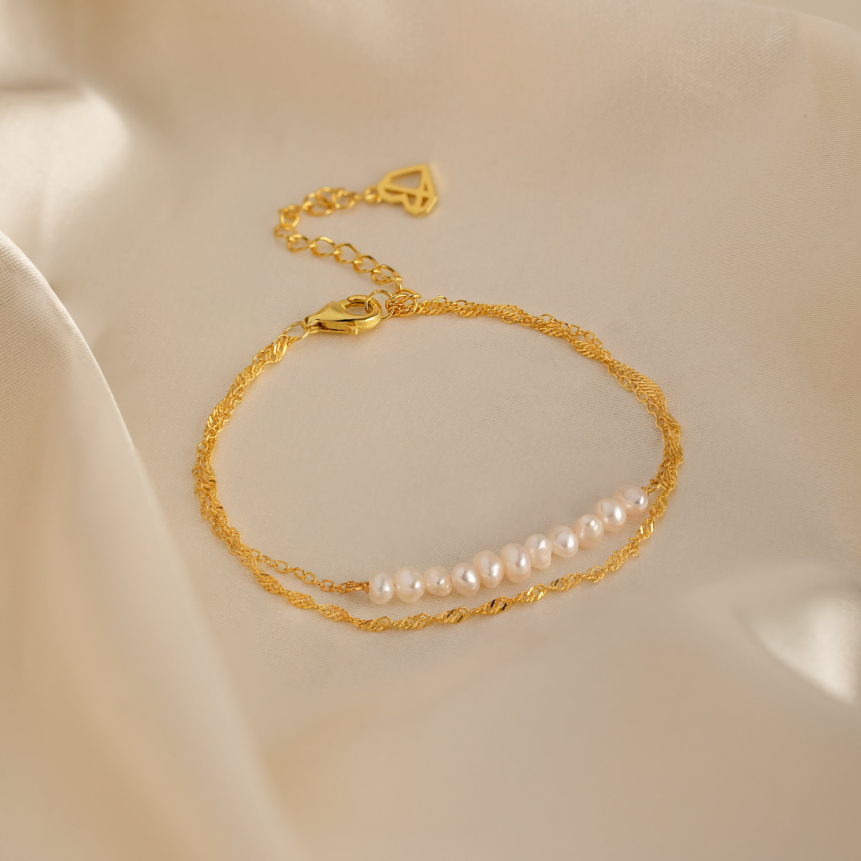 The Duo Pearl Twist Bracelet, featuring gold accents and white pearls, is elegantly displayed on soft beige satin fabric.