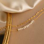 The Duo Pearl Twist Bracelet, featuring elegant pearls on a gold chain, rests on a gold-edged mirror next to soft, cream-colored fabric.