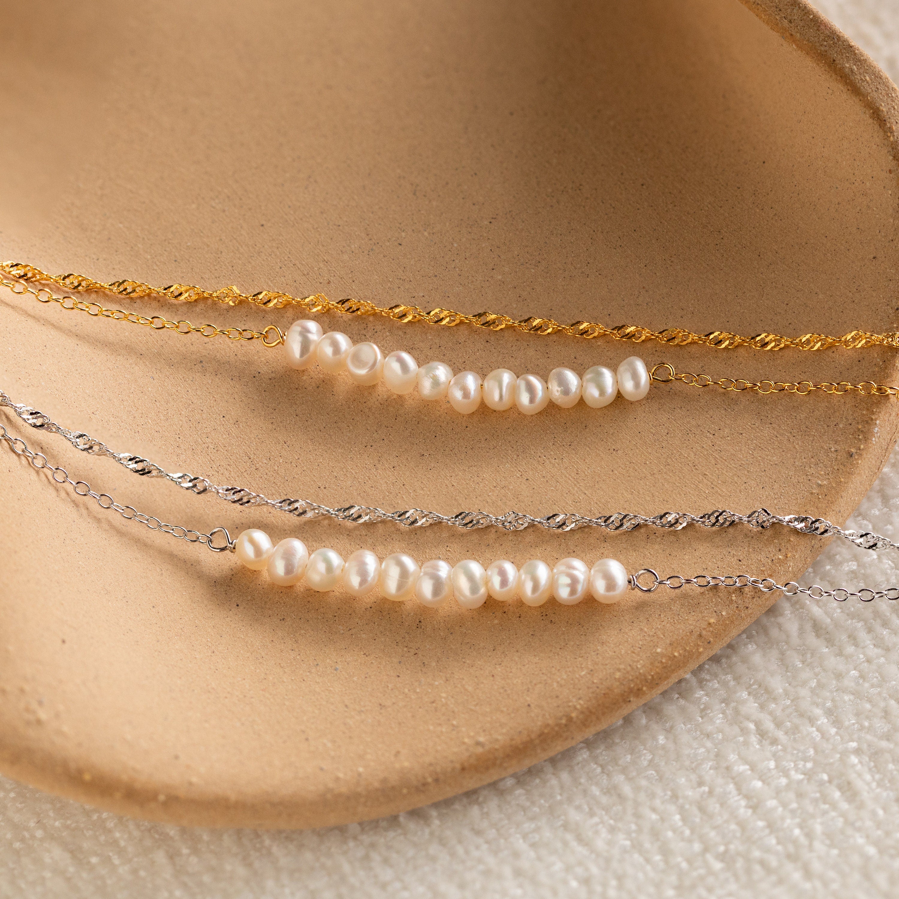 The Duo Pearl Twist Bracelet, along with two other delicate minimalist bracelets, is displayed on a curved beige ceramic dish.