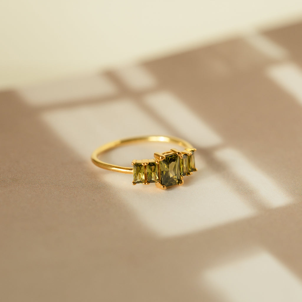 Art Deco Birthstone Ring—gold band with three baguette-cut green stones, displayed on a beige surface with soft shadows, radiating vintage charm.