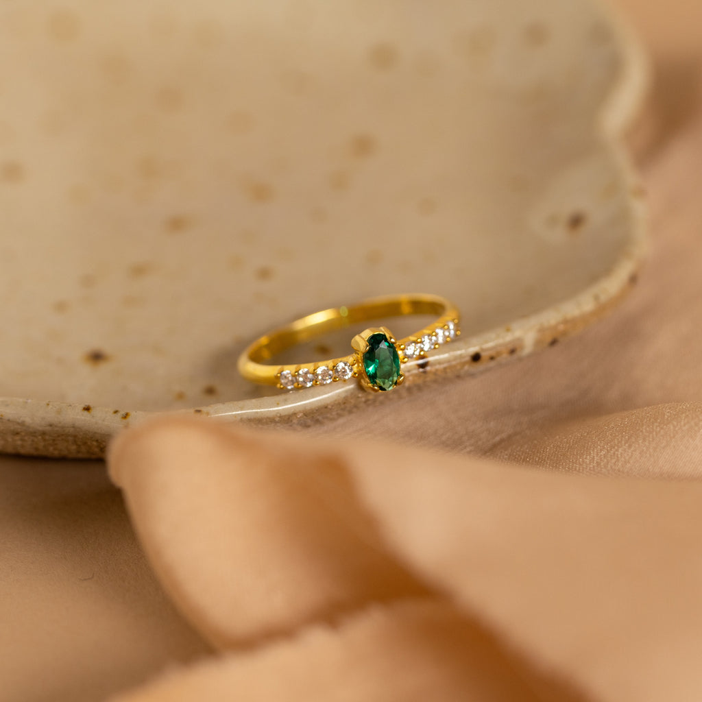 The Pave Oval Birthstone Ring, featuring a green oval center stone and pave diamonds, rests on a ceramic dish with beige fabric beside it.