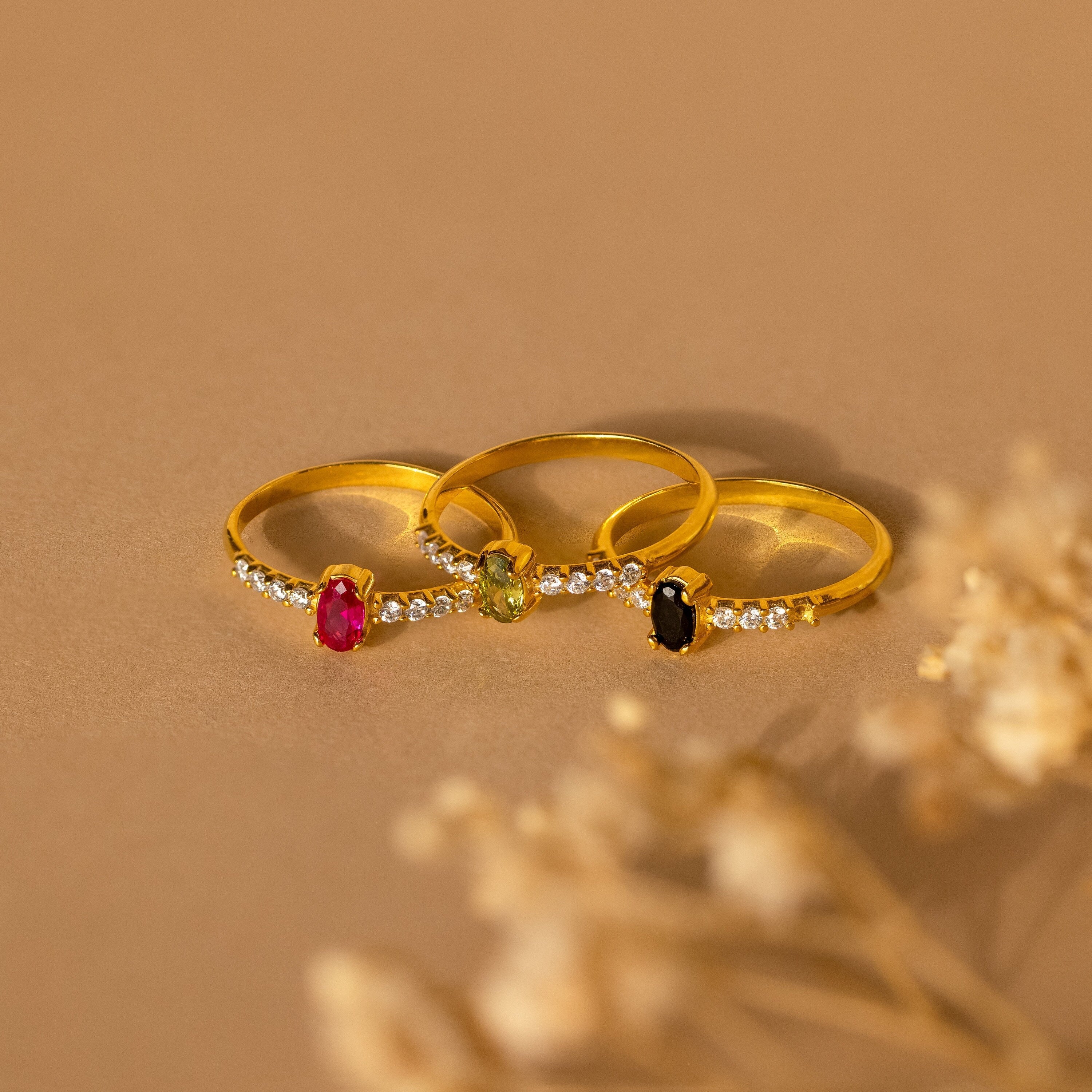 The Pave Oval Birthstone Ring, shown with sparkling gemstones and petite diamonds on a beige surface with dried flowers, is a stunning example of elegant gemstone ring design.