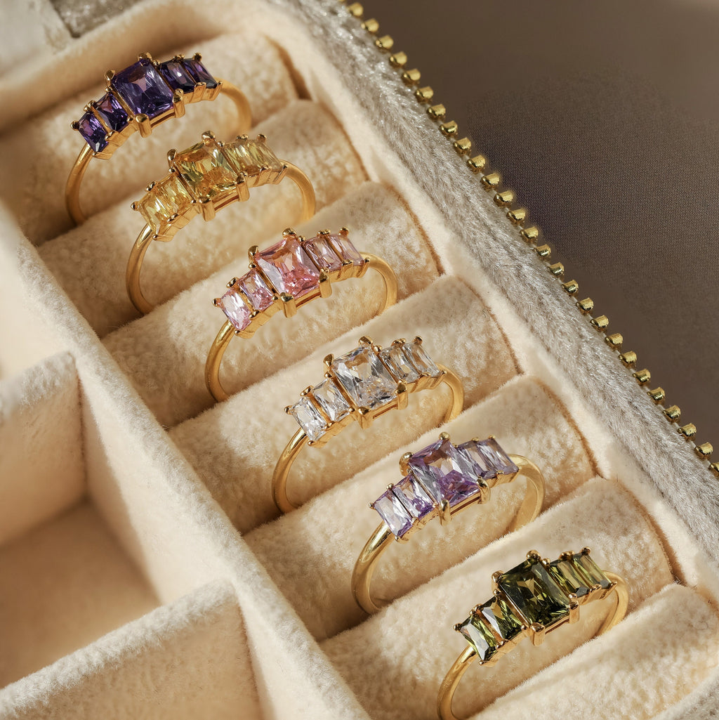 Six Art Deco Birthstone Rings, each featuring a colorful baguette-cut stone and vintage-inspired design, displayed elegantly in a beige jewelry box.