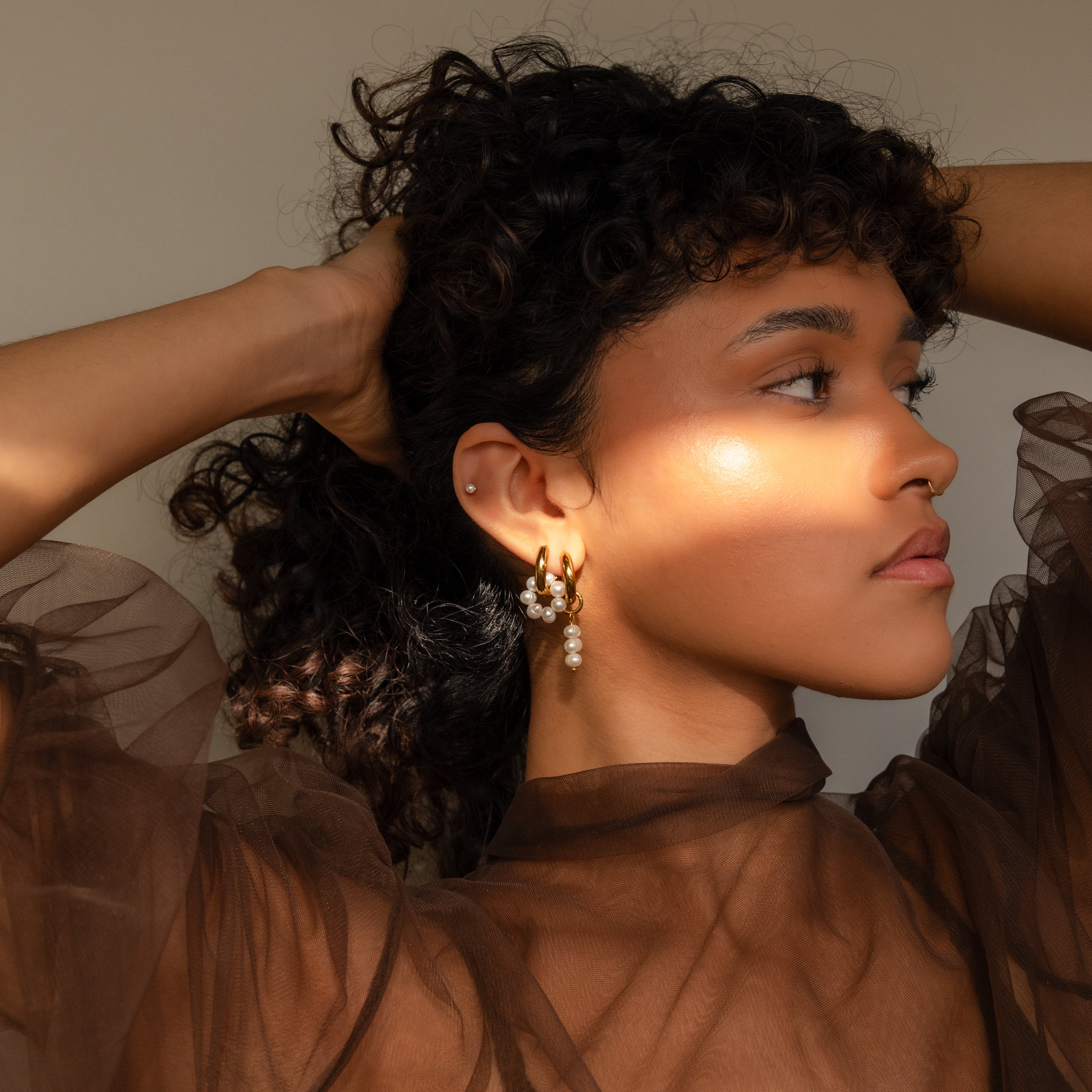 A woman with curly hair wears a sheer brown top and looks to the side in sunlight, showcasing mismatched pearl drop hoops.