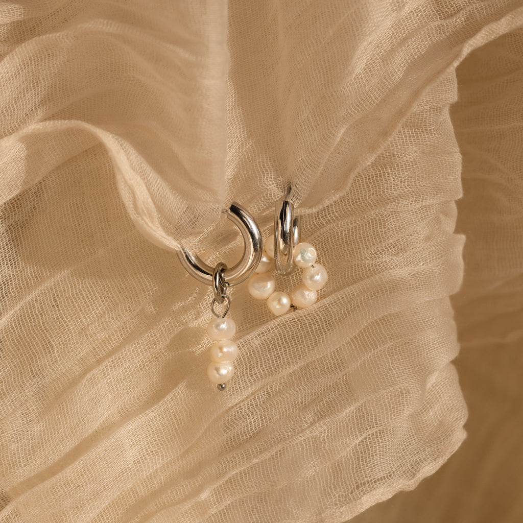 The Mismatched Pearl Drop Hoops feature two silver statement hoops adorned with small pearls, displayed on a textured beige fabric background—ideal as an elegant bridesmaid gift.