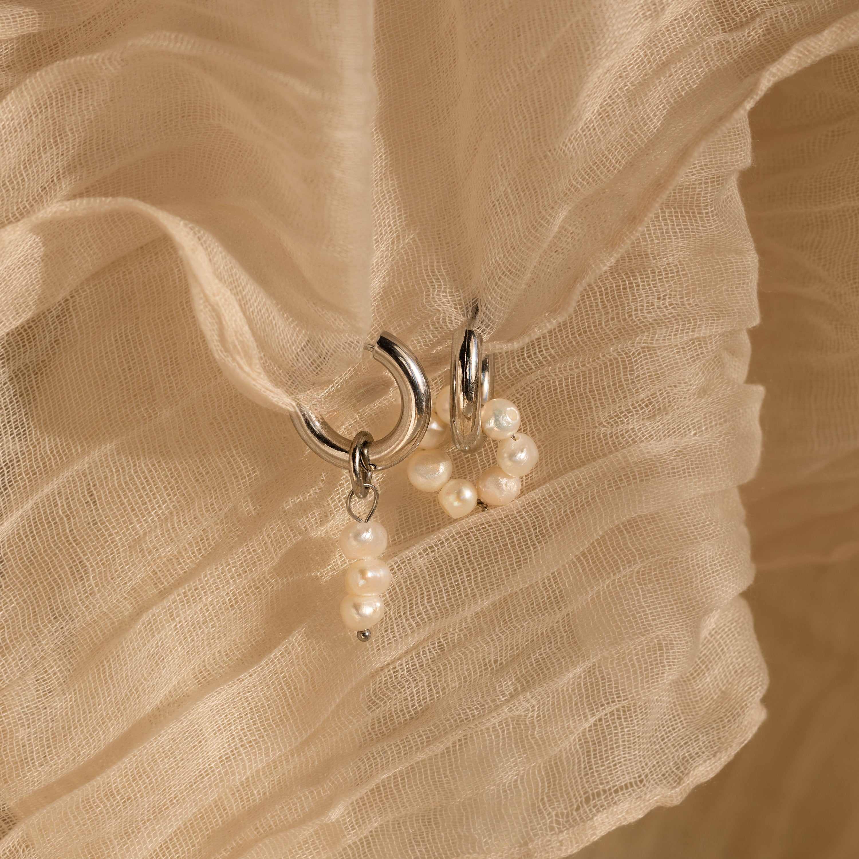 The Mismatched Pearl Drop Hoops feature two silver statement hoops adorned with small pearls, displayed on a textured beige fabric background—ideal as an elegant bridesmaid gift.