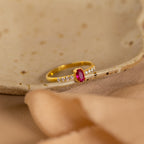The Pave Oval Birthstone Ring features a red center stone accented with small clear stones, displayed on a ceramic dish with a soft beige cloth. This elegant custom piece brings sophistication to any setting.