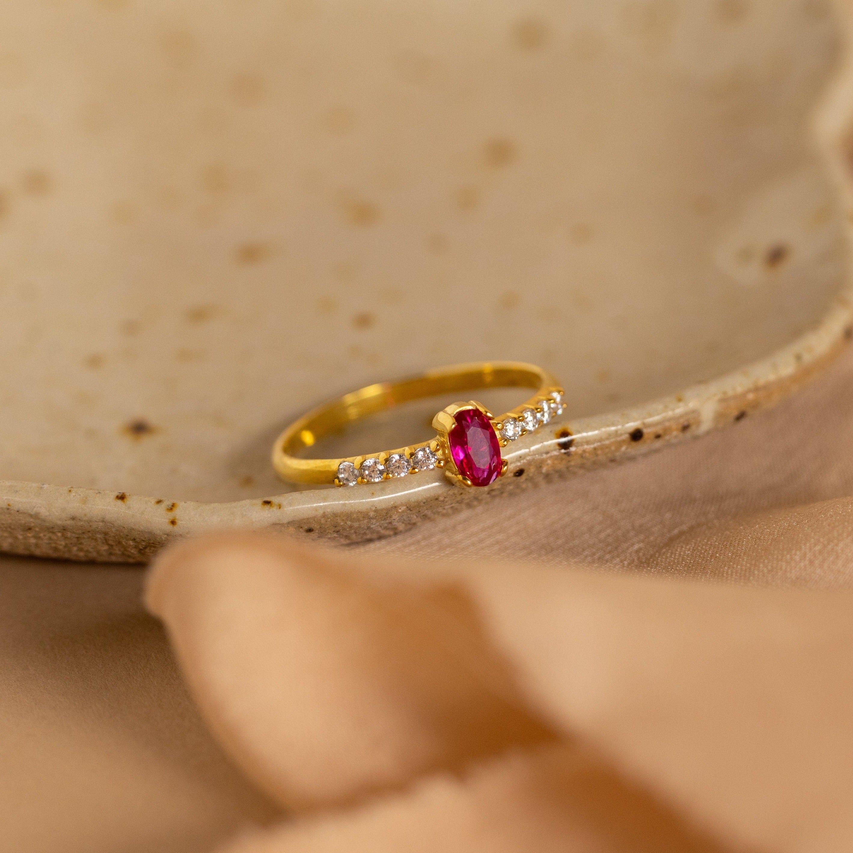 The Pave Oval Birthstone Ring features a red center stone accented with small clear stones, displayed on a ceramic dish with a soft beige cloth. This elegant custom piece brings sophistication to any setting.
