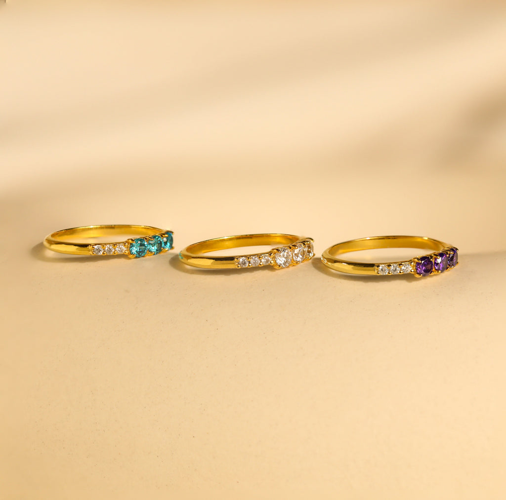 The Pave Triple Birthstone Ring features three gold bands with blue, white, and purple gemstones on a beige surface—ideal for anyone seeking a customizable birthstone ring and unique personalized jewelry.