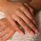 A hand with pink nails wears the Pave Princess Birthstone Ring, featuring a gold band, red gemstone, and small diamonds, resting on a textured surface.
