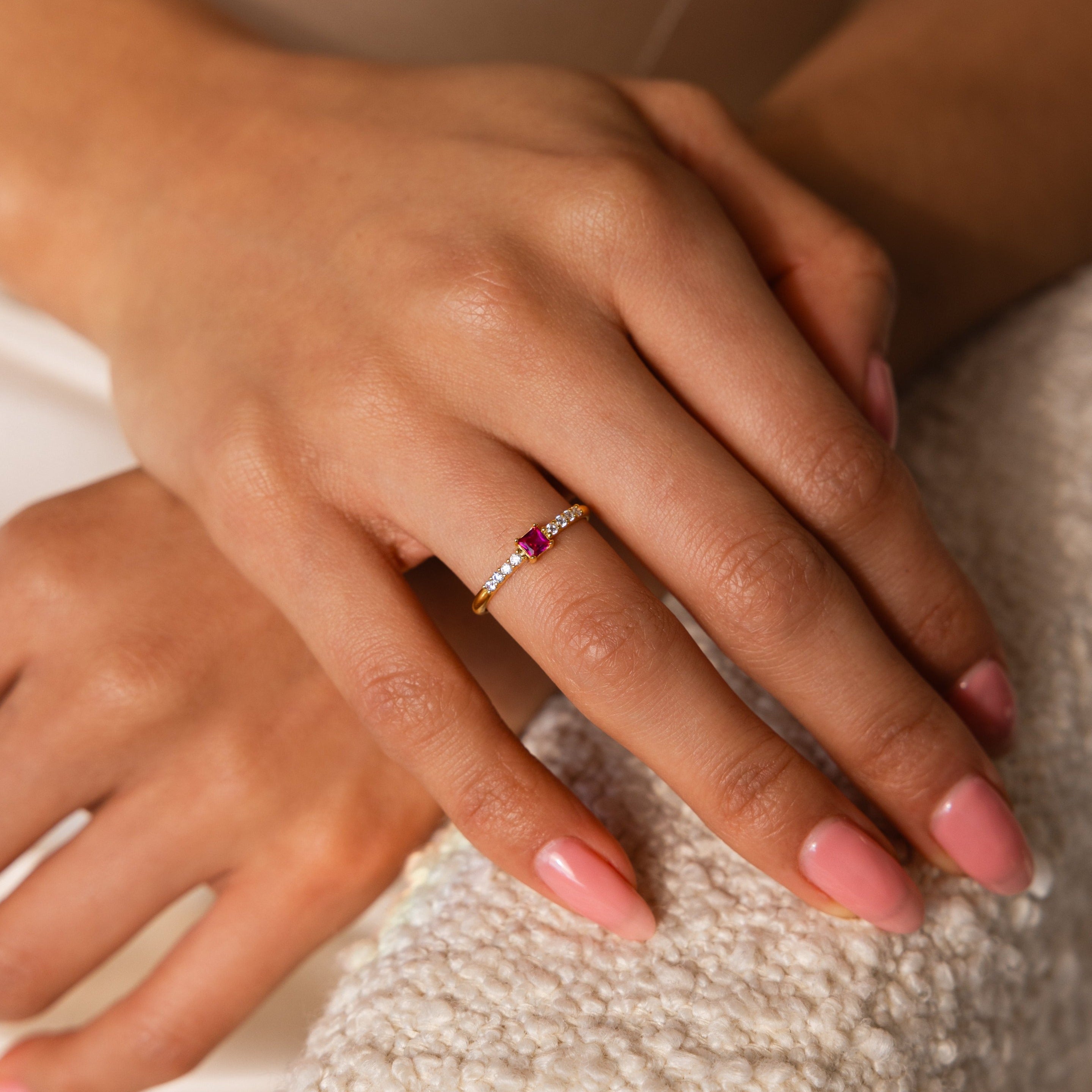 A hand with pink nails wears the Pave Princess Birthstone Ring, featuring a gold band, red gemstone, and small diamonds, resting on a textured surface.