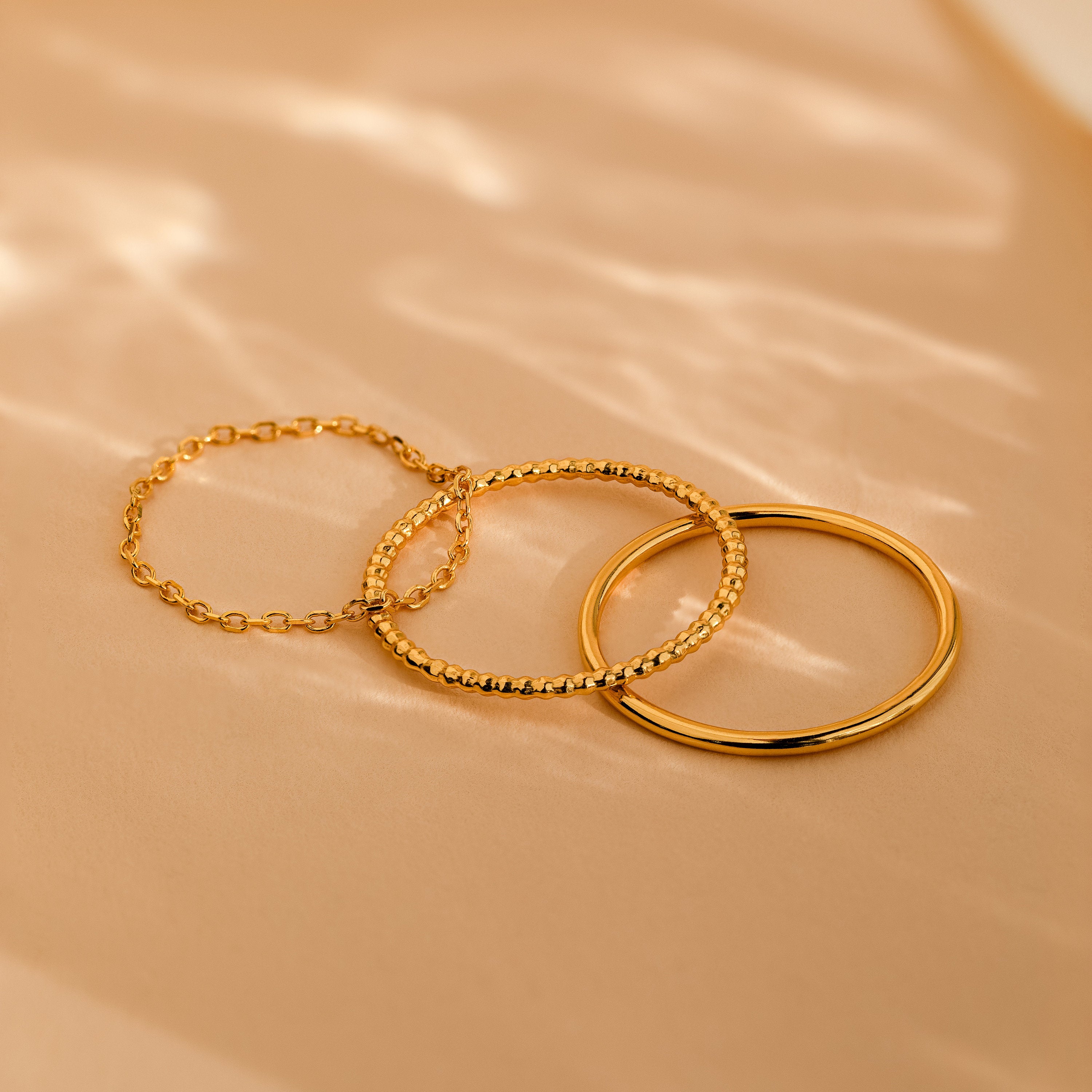 Three delicate rings from the Textured Stacking Ring Set, each featuring a unique texture, are arranged on a beige surface under soft natural lighting.