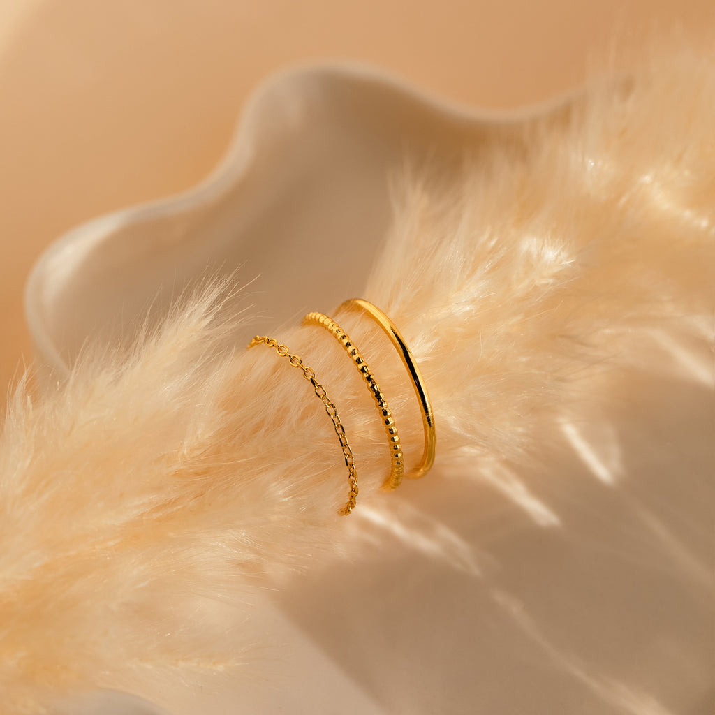 Three dainty rings from our Textured Stacking Ring Set are displayed on soft beige pampas grass, with a wavy white ceramic dish in the background—ideal for fans of stylish stacking jewelry.