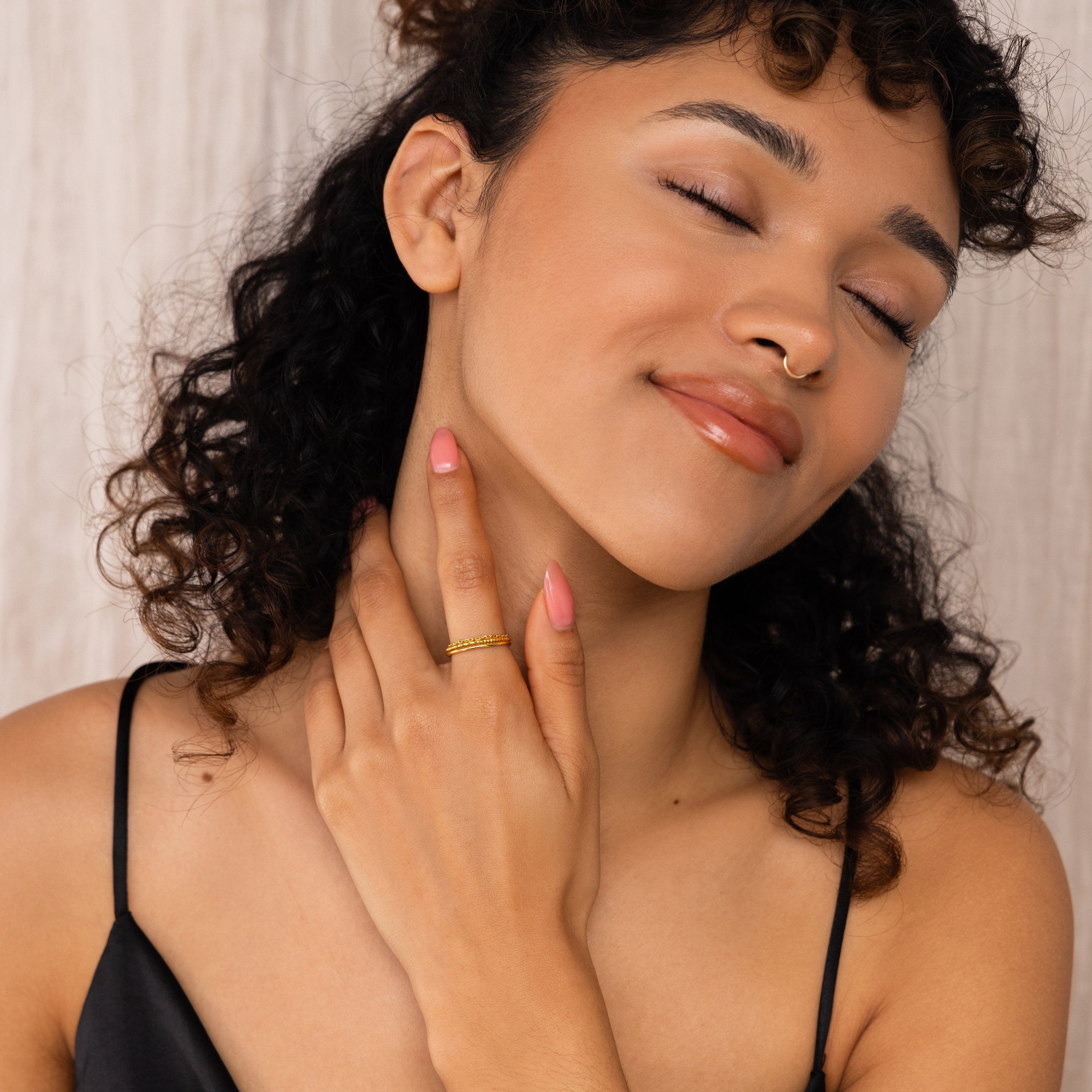 A woman with curly hair and closed eyes smiles gently, touching her neck while wearing a black top and the Textured Stacking Ring Set in gold.