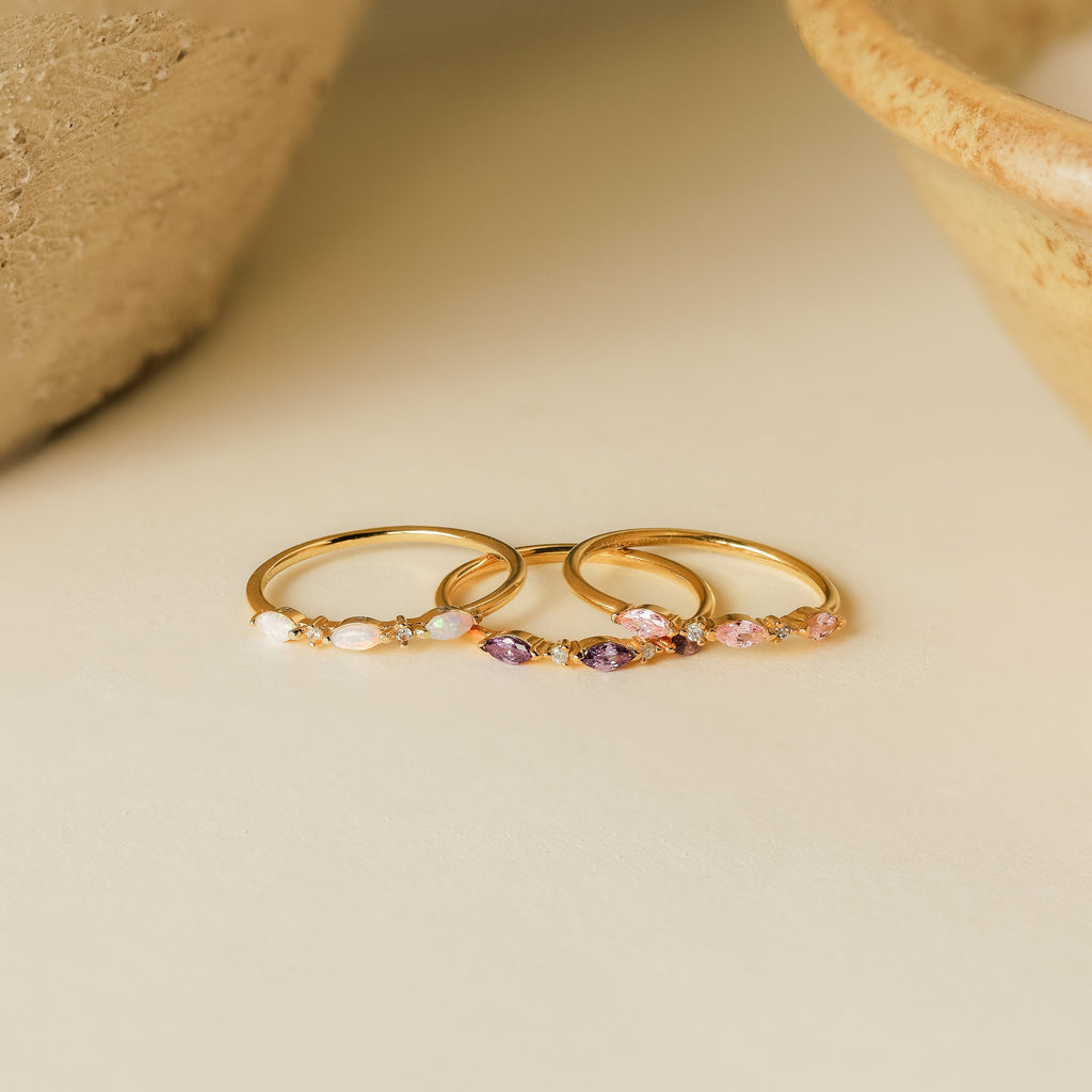 The Triple Marquise Birthstone Ring, a delicate gold stacking ring with small colored gemstones, including CZ Diamond, is displayed on a light neutral surface.