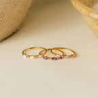 The Triple Marquise Birthstone Ring, a delicate gold stacking ring with small colored gemstones, including CZ Diamond, is displayed on a light neutral surface.