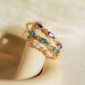 The Triple Marquise Birthstone Ring features three gold stacking bands with marquise-cut purple, blue, and clear CZ gemstones, elegantly arranged on a cream-colored surface.