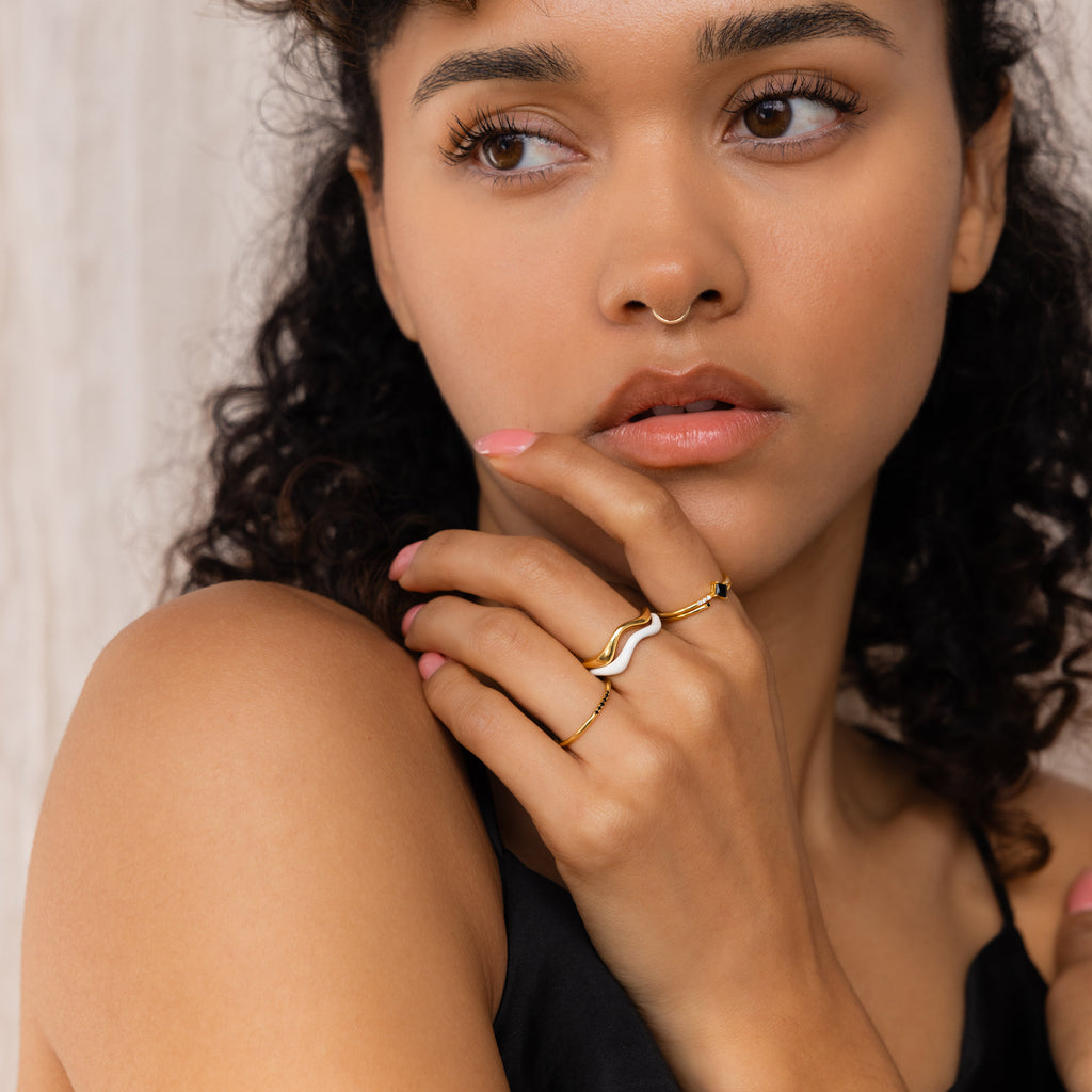 A woman with curly hair wears minimalist rings, featuring the Duo Wave Ring Set, and a nose ring while looking to the side with her hand near her lips.