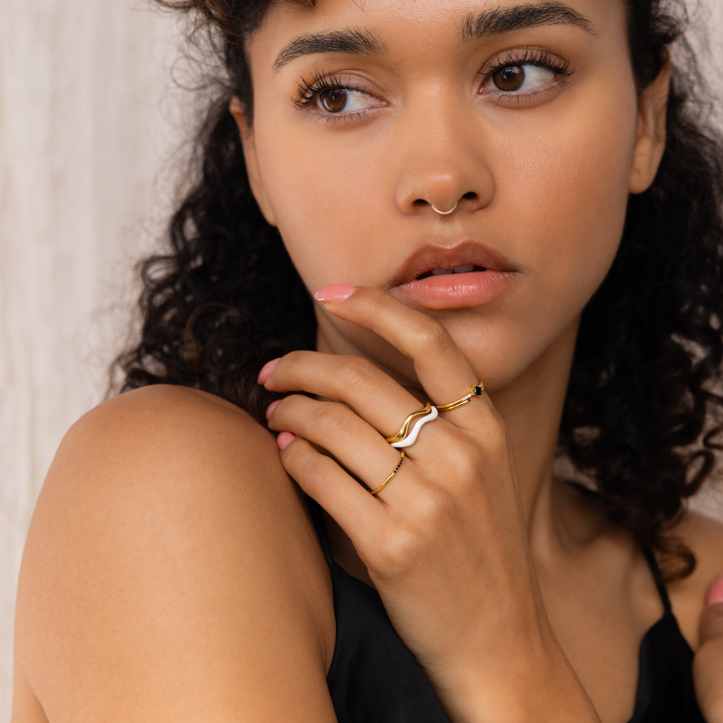 A woman with curly hair wears minimalist rings, featuring the Duo Wave Ring Set, and a nose ring while looking to the side with her hand near her lips.