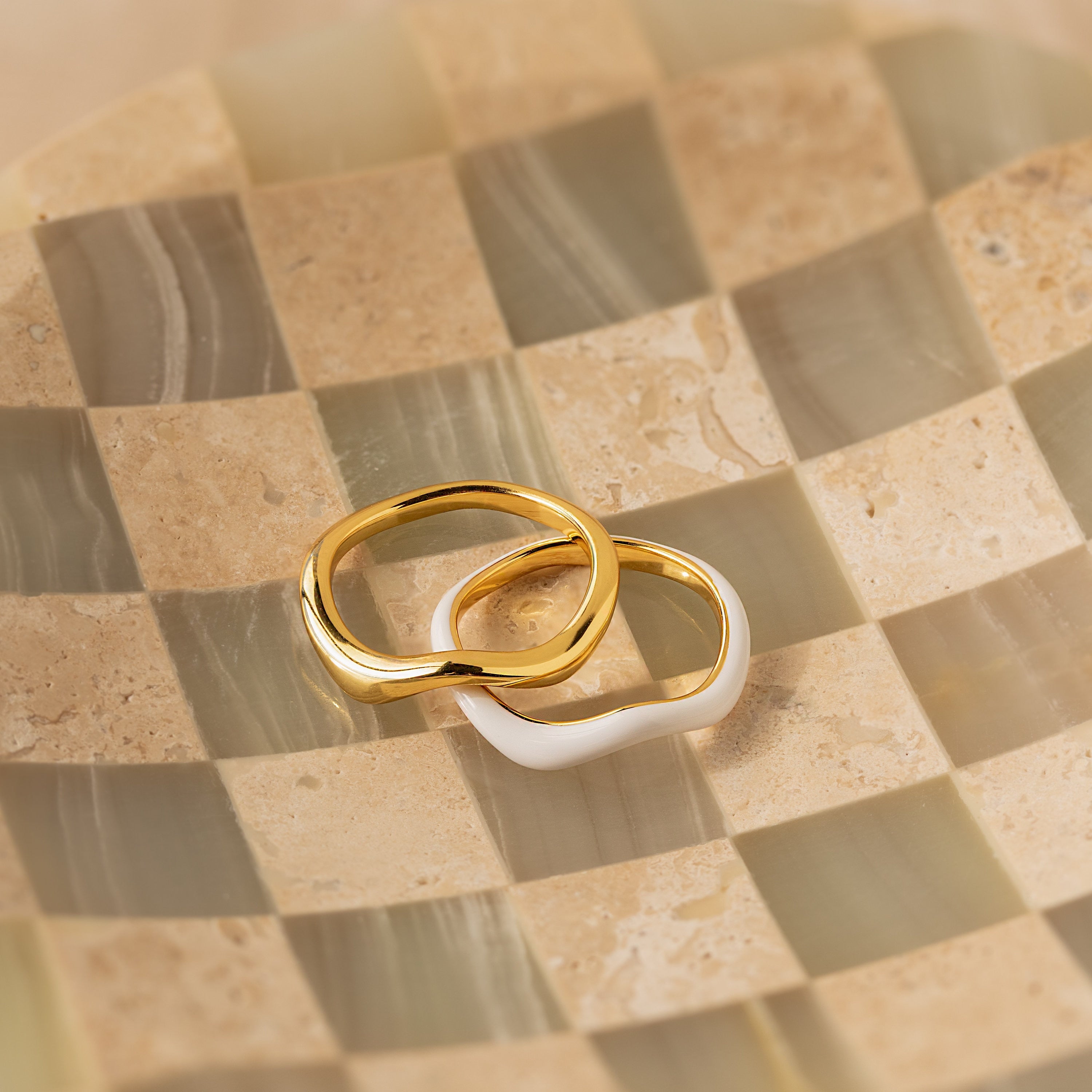 The Duo Wave Ring Set features two wavy rings, one gold and one silver, showcased on a glossy beige and cream checkerboard surface.