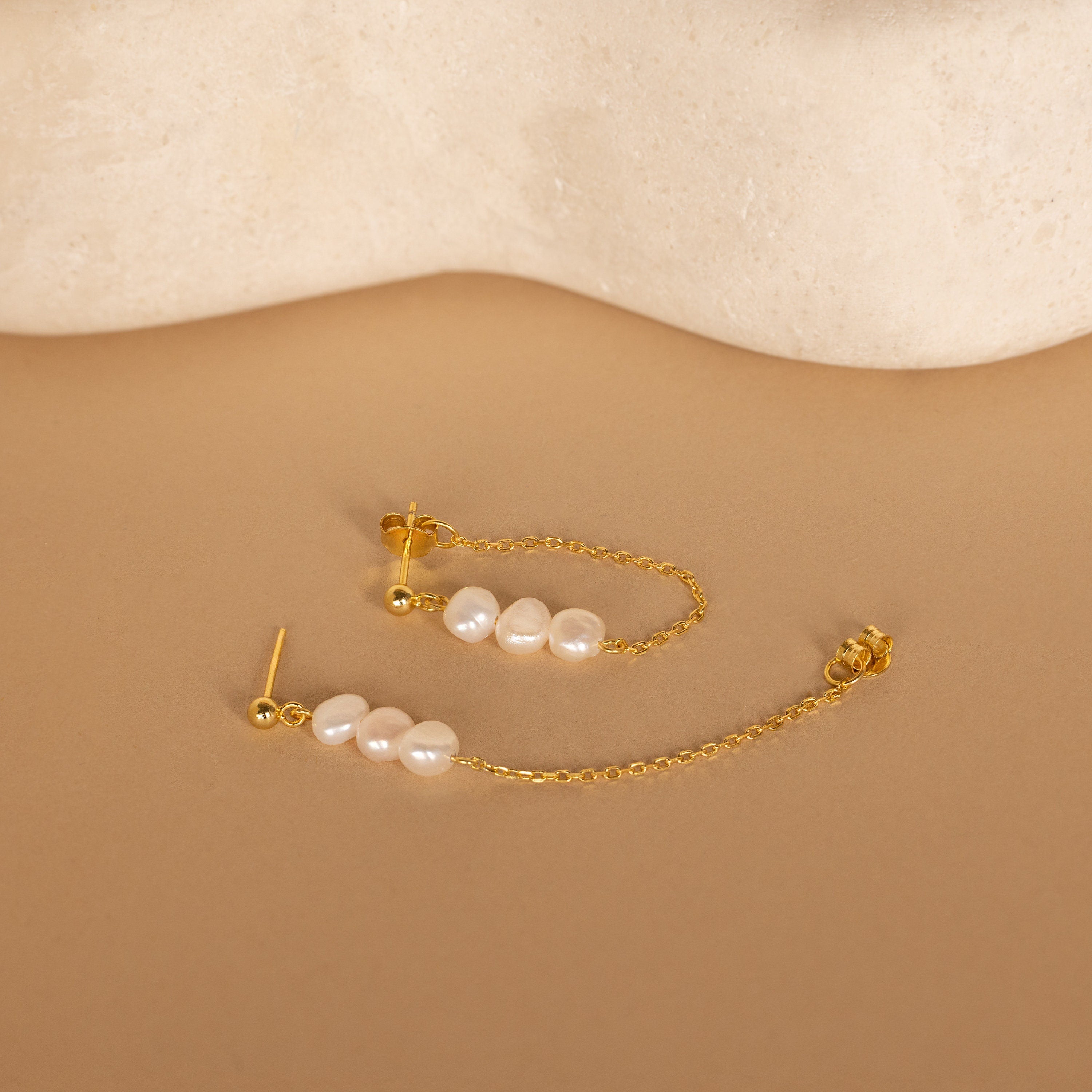 Triple Pearl Drop Earrings feature gold chains and small white pearls, beautifully displayed on a beige surface with a stone background.