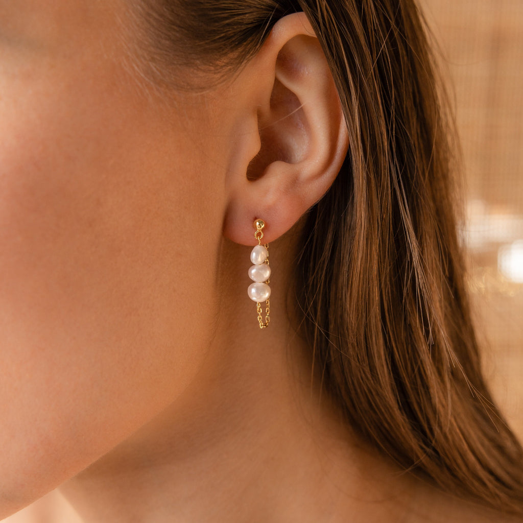 Close-up of a woman's ear wearing Triple Pearl Drop Earrings, showcasing three white pearls in a gold setting—a timeless and elegant jewelry accessory.