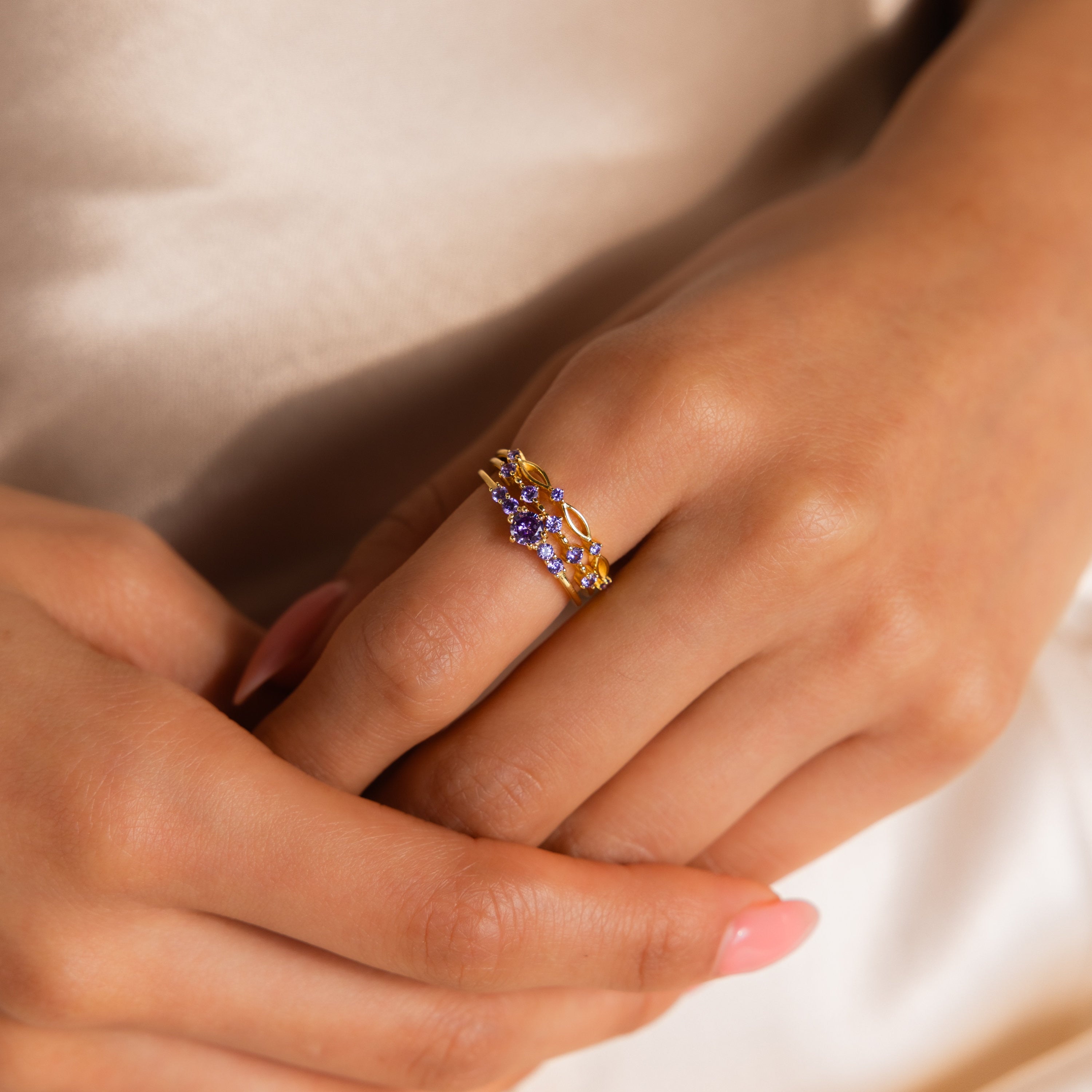 A hand wearing the Amethyst Stacking Ring Set—gold bands with purple gemstone and clear accents, fingers gently intertwined; a chic choice for those who love fashion-forward jewelry.