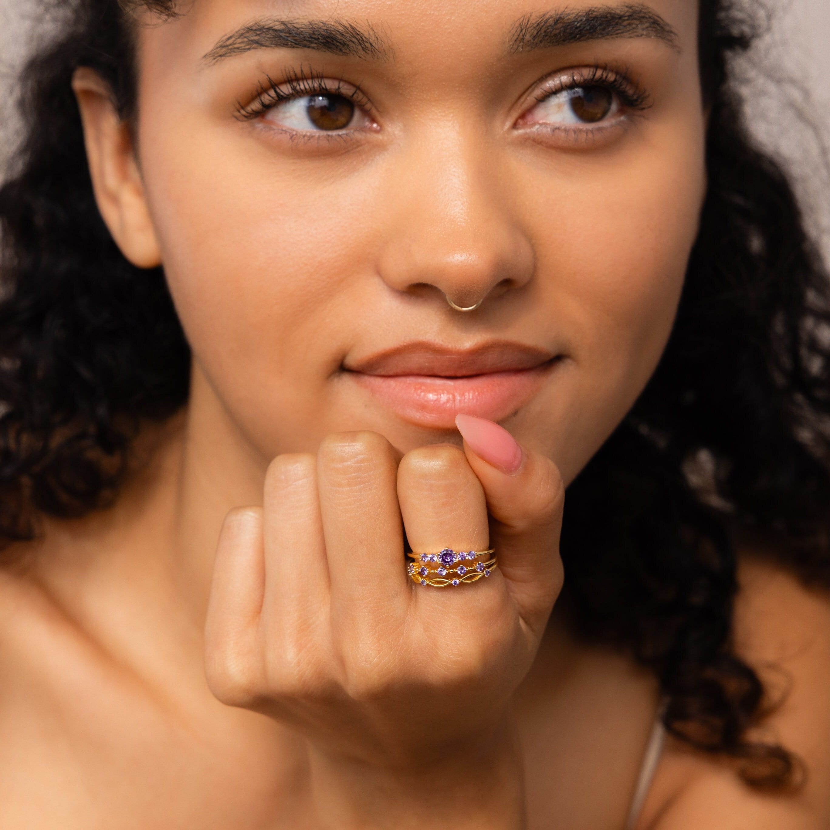 A woman with curly hair, looking thoughtful, rests her chin on her hand while wearing the Amethyst Stacking Ring Set featuring gold bands.