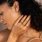 A woman with curly hair touches her neck, wearing gold stacking rings and the Mini Signet Mood Ring on her finger.