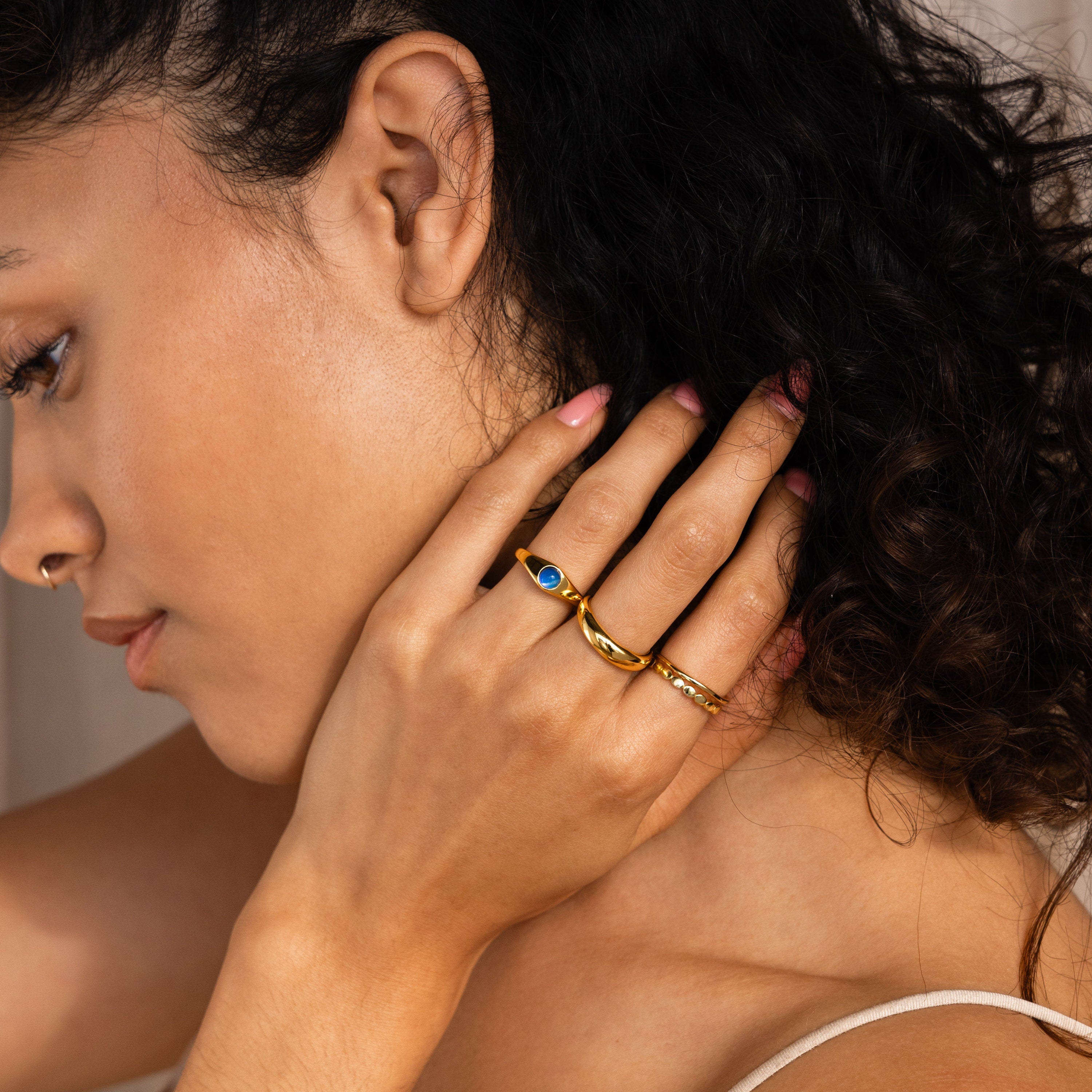 A woman with curly hair touches her neck, wearing gold stacking rings and the Mini Signet Mood Ring on her finger.