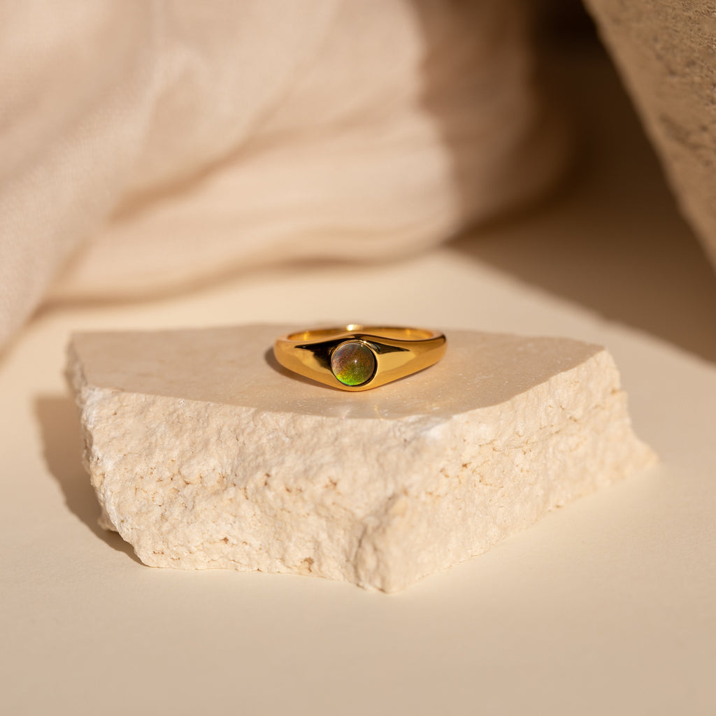 The Mini Signet Mood Ring is a minimalist gold ring with a green gemstone, displayed on a textured beige stone—ideal for stacking ring lovers.