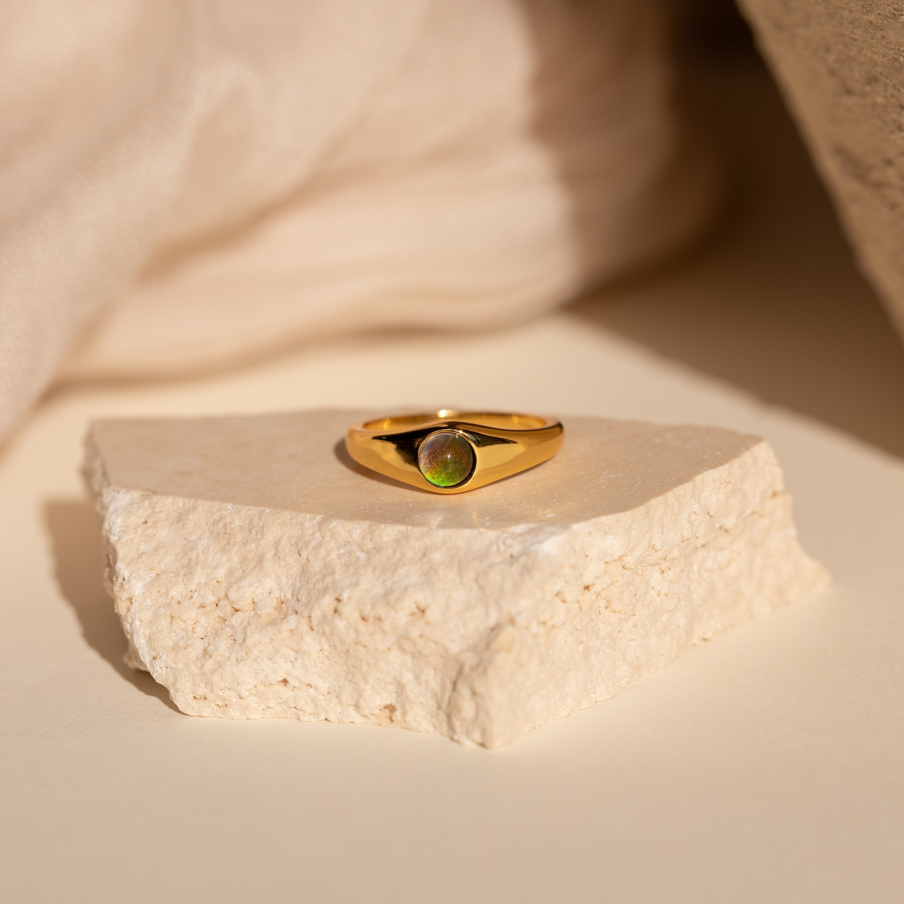 The Mini Signet Mood Ring is a minimalist gold ring with a green gemstone, displayed on a textured beige stone—ideal for stacking ring lovers.