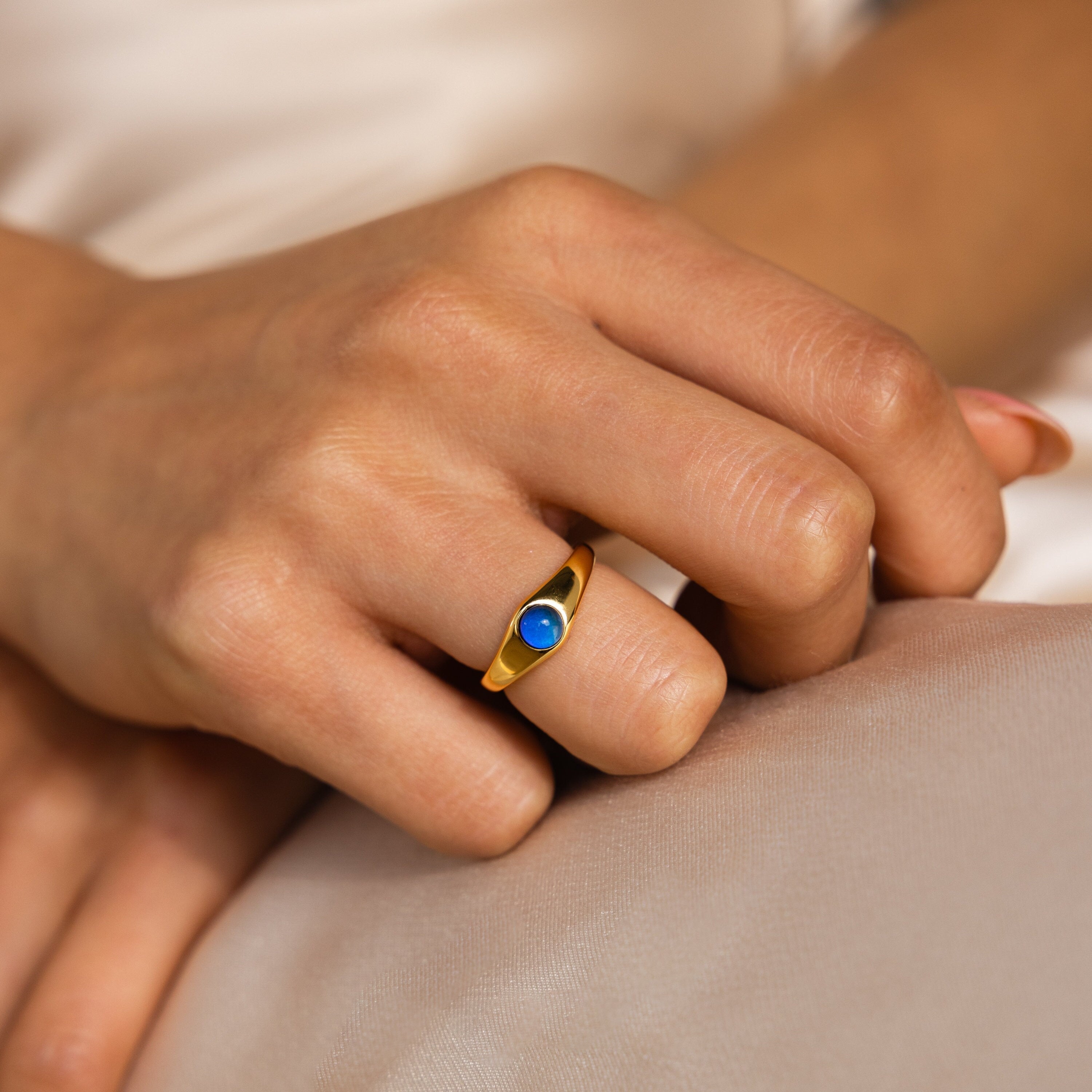 A hand wears the Mini Signet Mood Ring—a minimalist gold ring with a small round blue stone, ideal for stacking—resting on a light fabric surface.