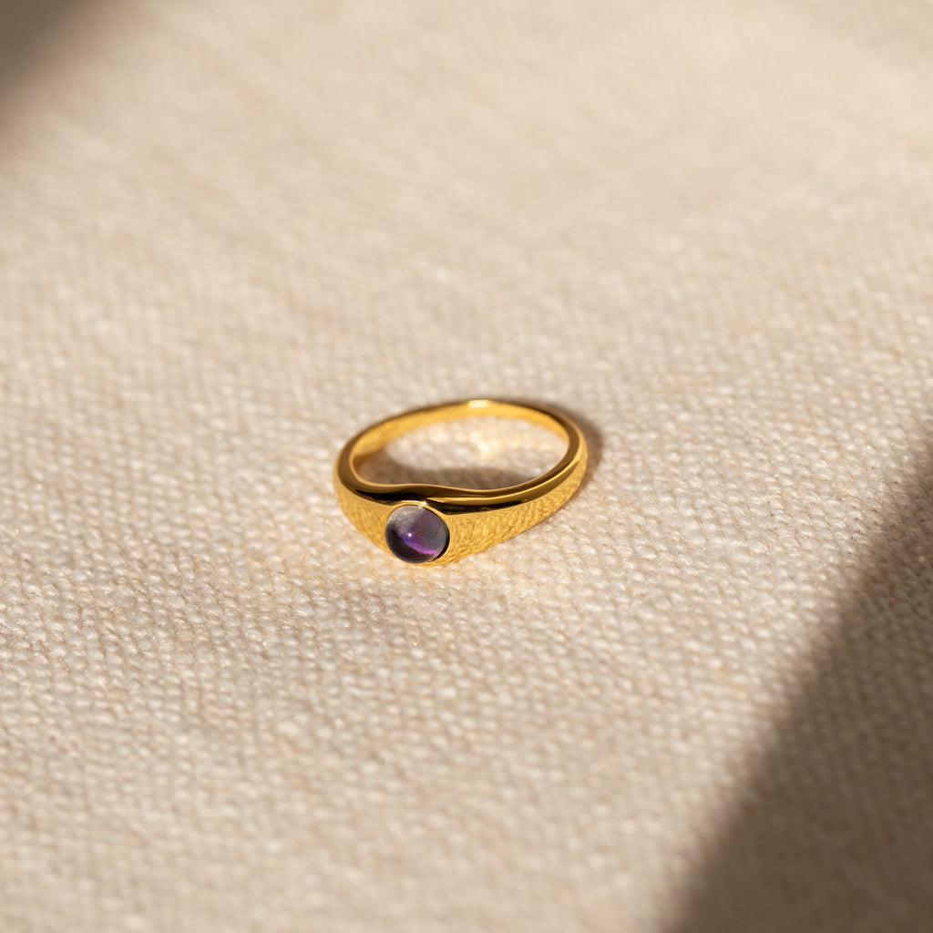 The Mini Signet Mood Ring is a minimalist gold band with a small purple gemstone, displayed on a textured beige surface in soft light—ideal for stacking or wearing as a chic statement piece.