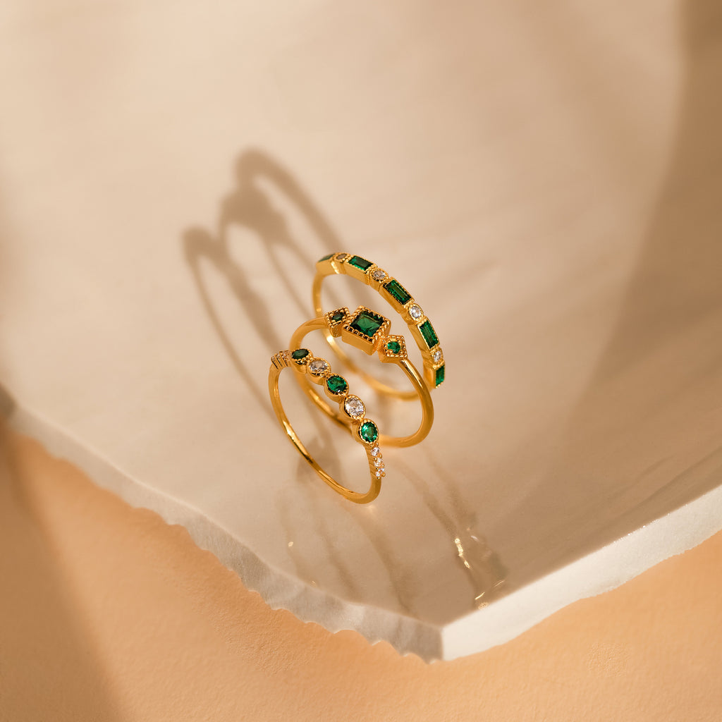 The Emerald Stacking Ring Set features three gold rings with green and white gemstones, displayed on a light textured surface. Ideal for lovers of Art Deco jewelry and vintage-inspired elegance.