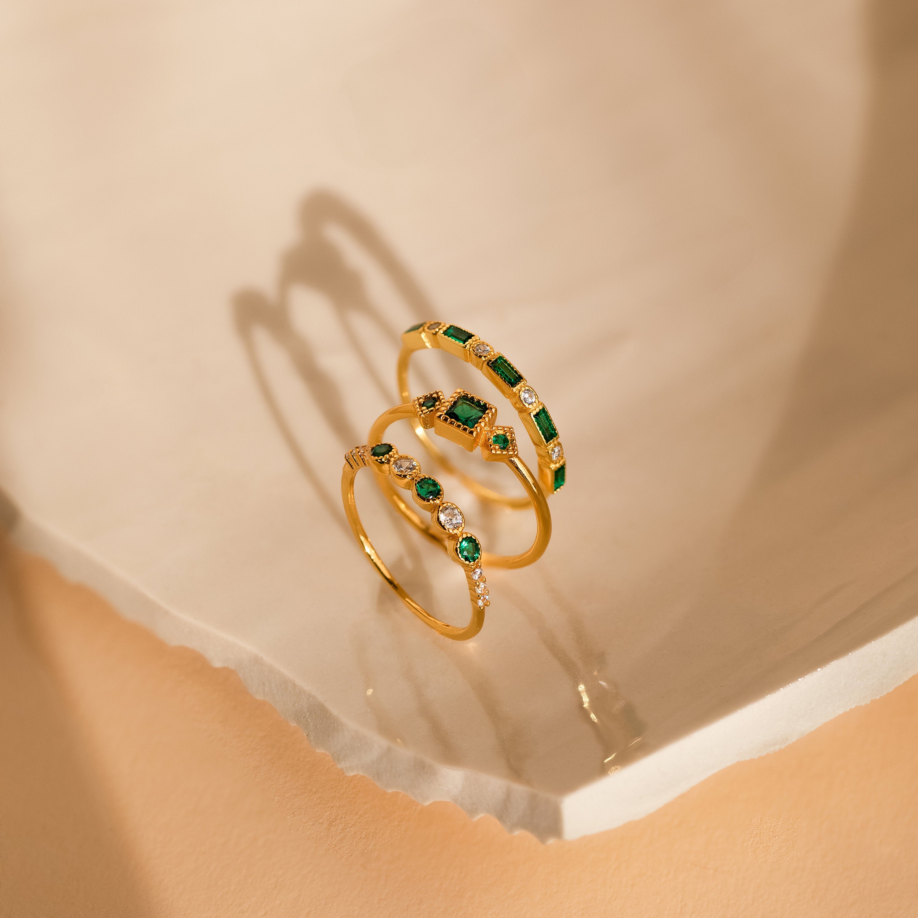 The Emerald Stacking Ring Set features three gold rings with green and white gemstones, displayed on a light textured surface. Ideal for lovers of Art Deco jewelry and vintage-inspired elegance.