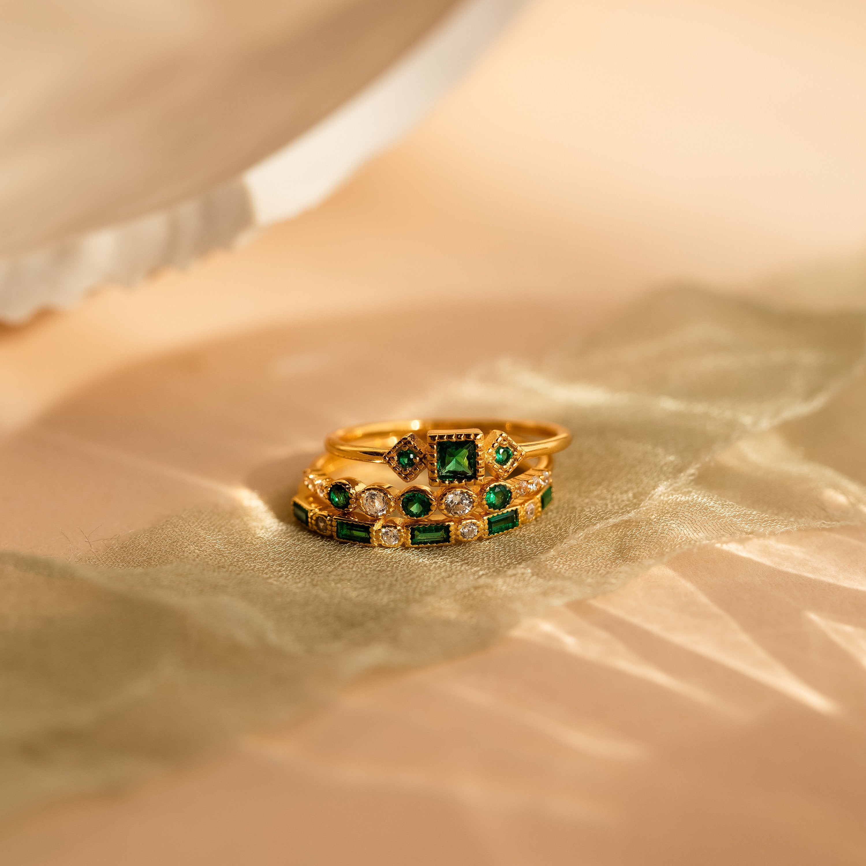 The Emerald Stacking Ring Set in 18K Gold features vintage-inspired gold rings with emerald accents and sparkling diamonds, displayed on soft green fabric that highlights their art deco elegance.