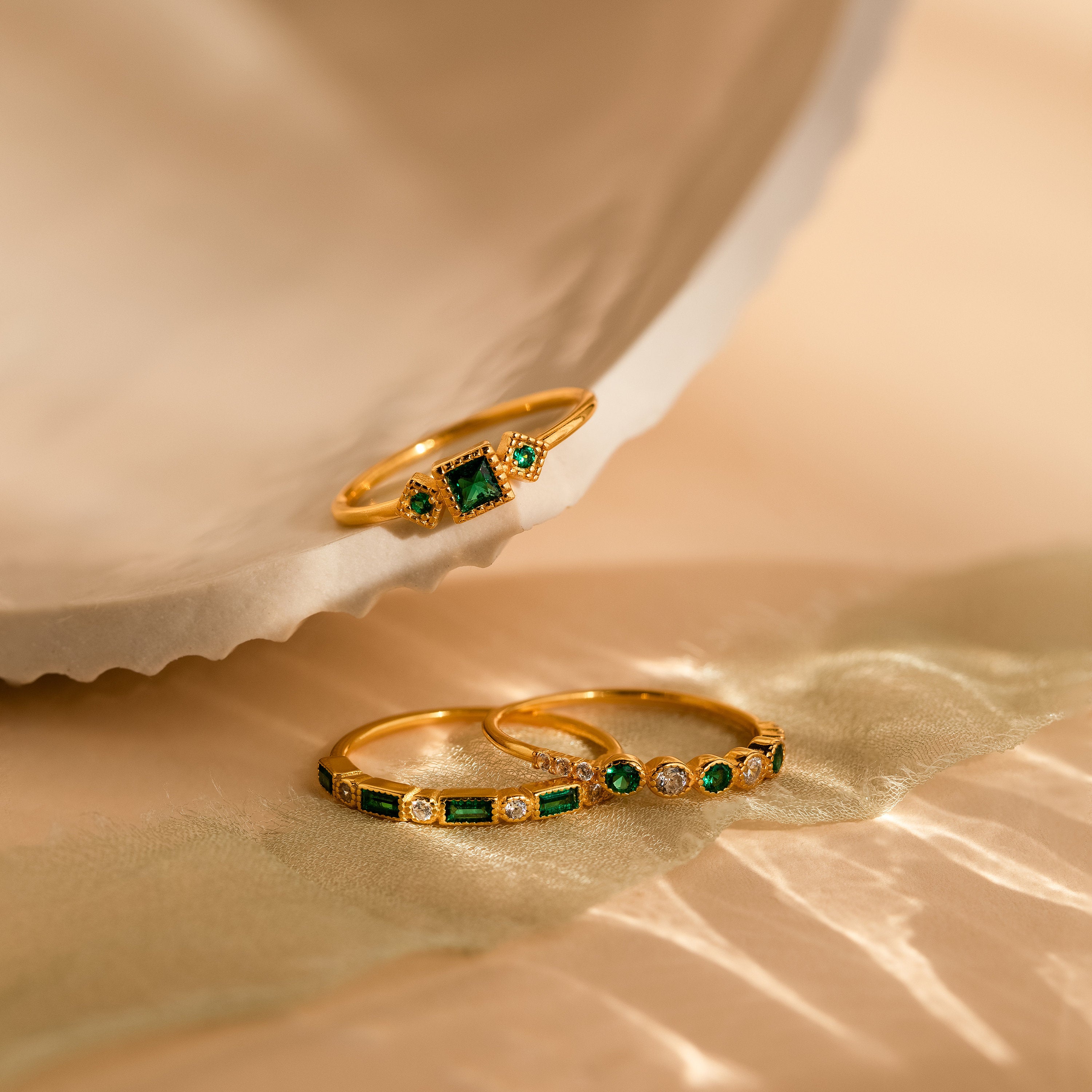 The Emerald Stacking Ring Set in 18K Gold is displayed on a shell and soft, light fabric surface alongside two other gold rings, one of which is vintage-inspired with green and white gemstones.